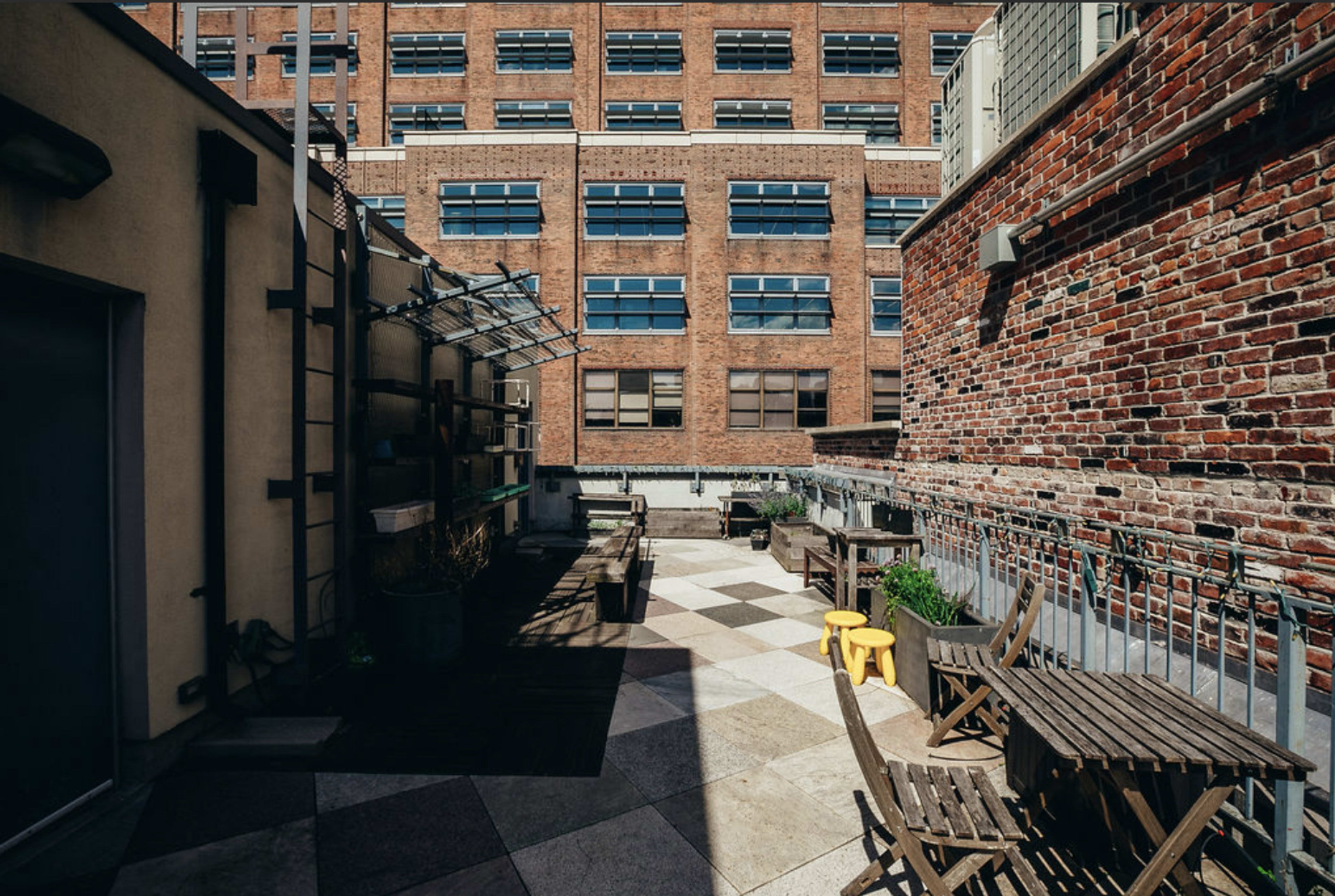 The image shows a rooftop terrace with wooden furniture, planters, and a view of a brick building with large windows.