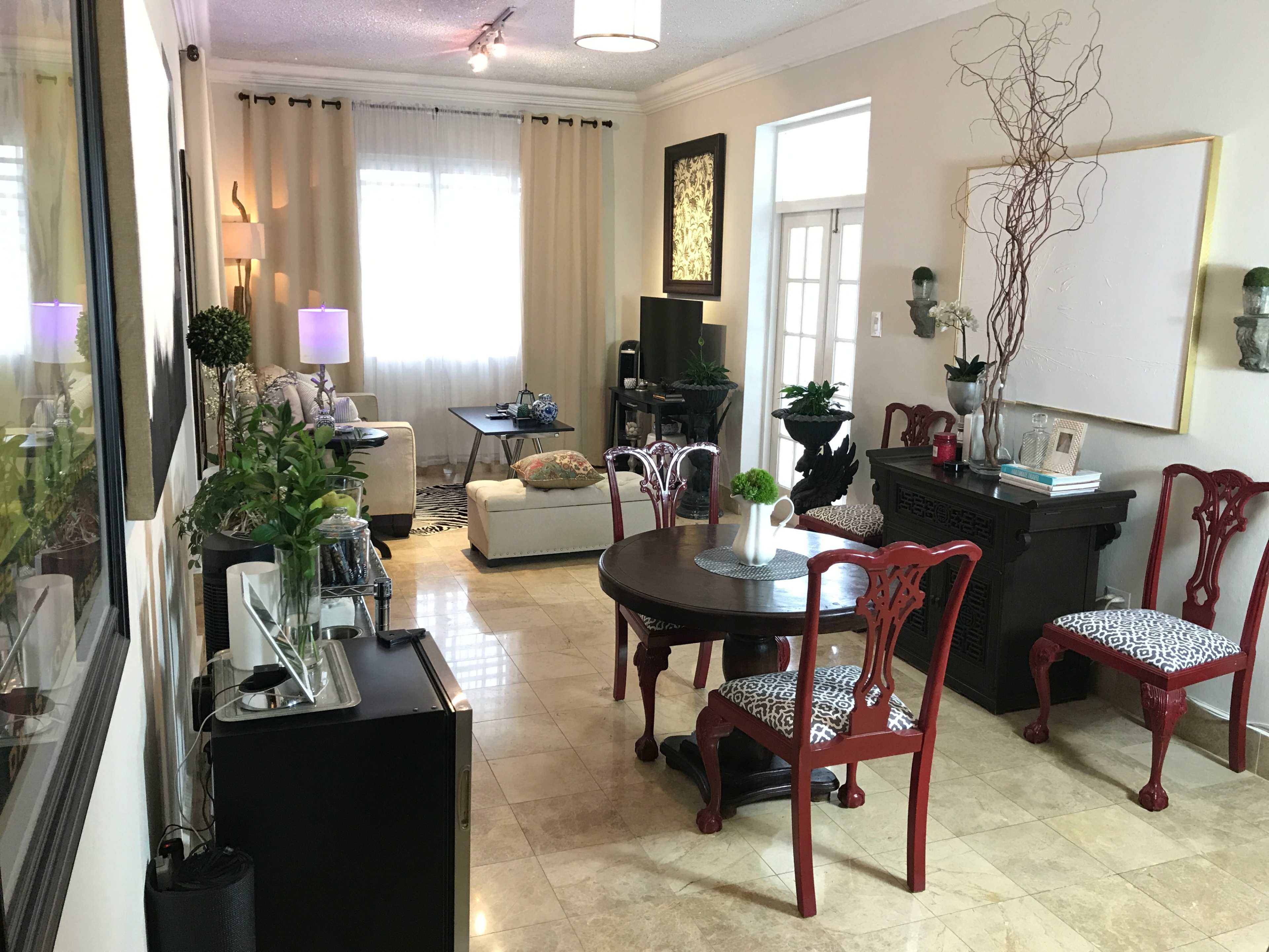 The image shows a living room with a dining table, red chairs, potted plants, and light-colored decor.