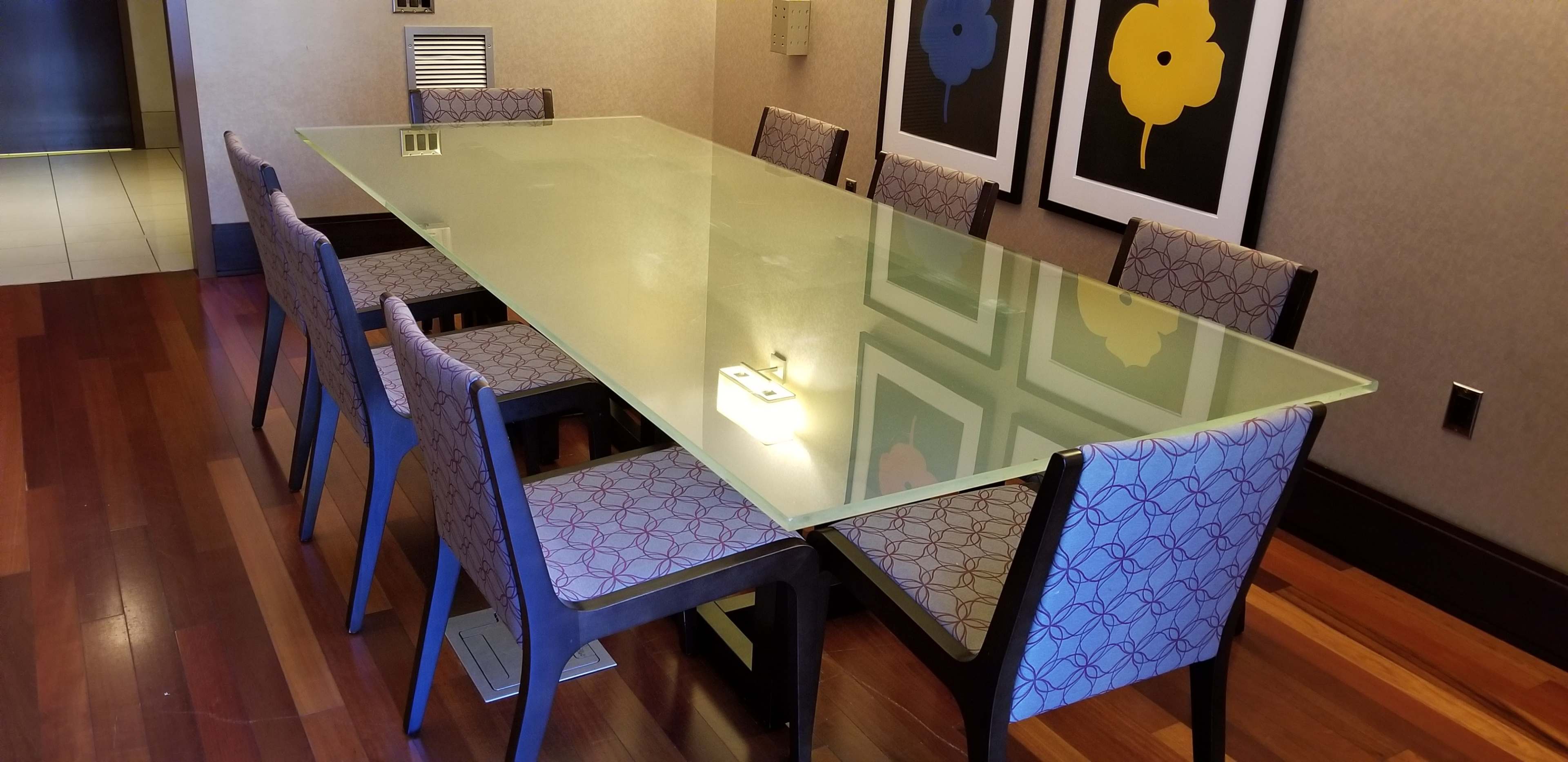 A long glass dining table with eight patterned chairs is surrounded by neutral walls and framed floral artwork.