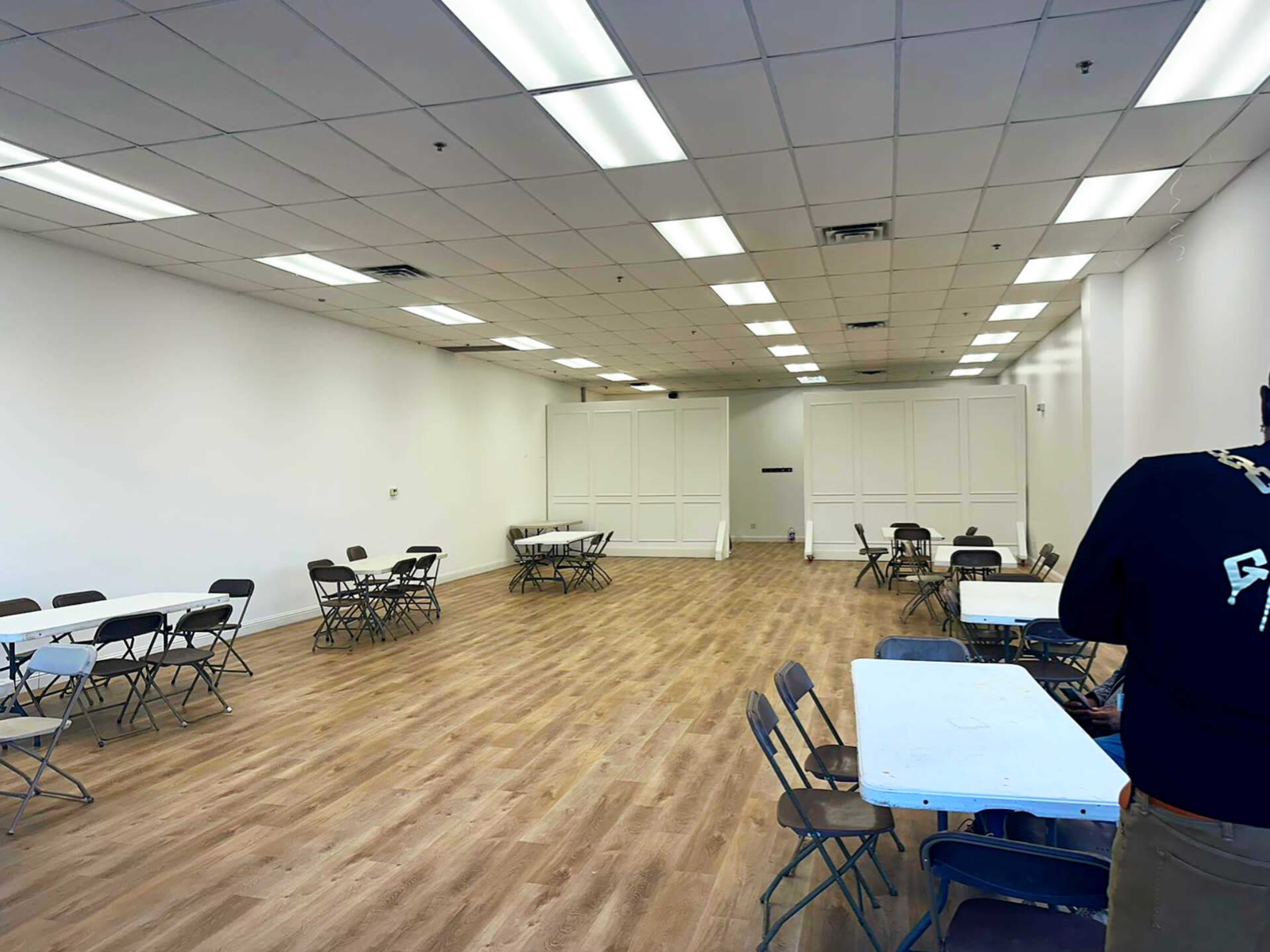 A spacious, empty room with a light-colored wooden floor and several folding tables and chairs arranged throughout.