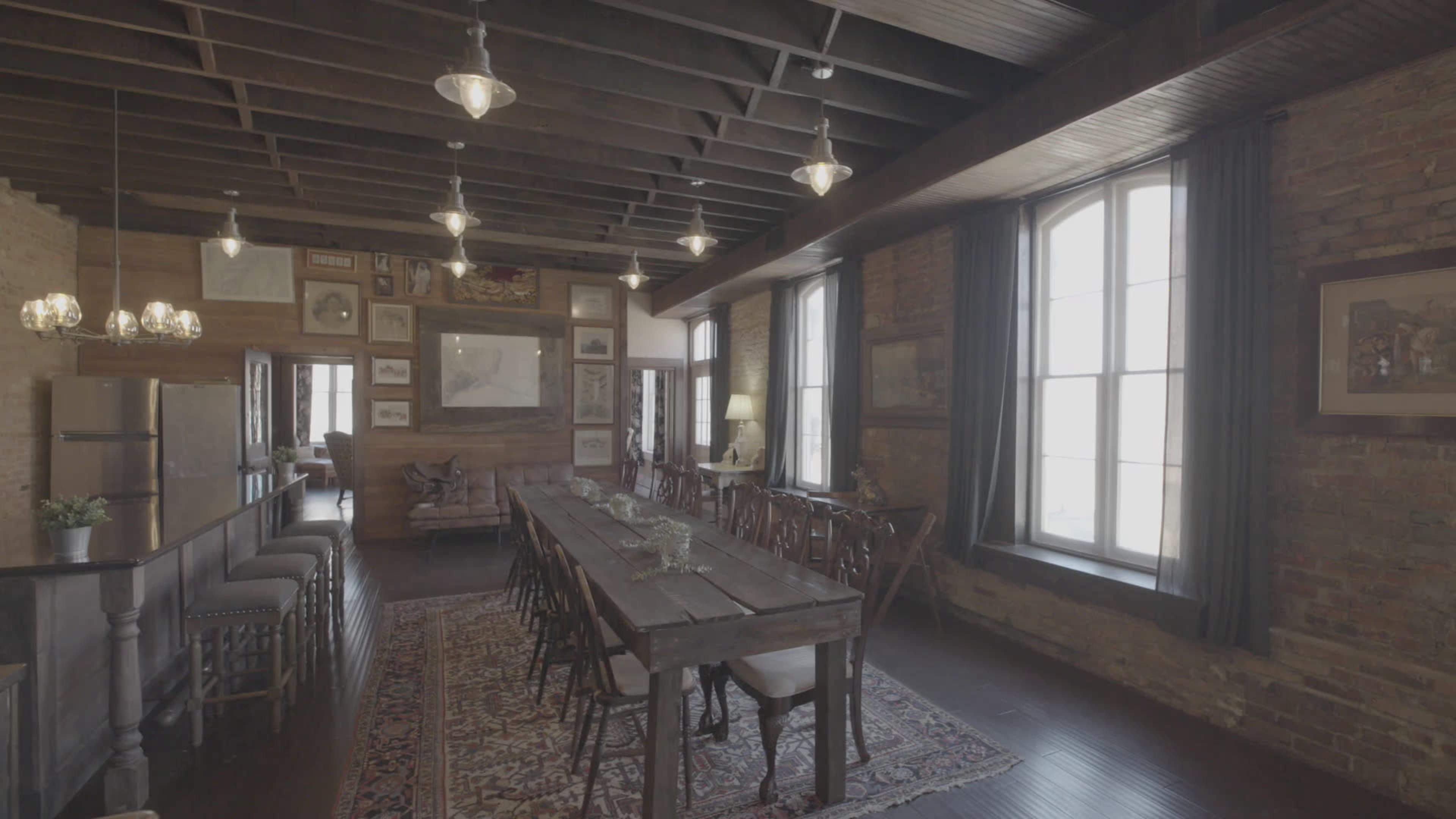 A spacious dining room features a long wooden table surrounded by chairs, with large windows allowing natural light to illuminate the rustic decor on the exposed brick walls.