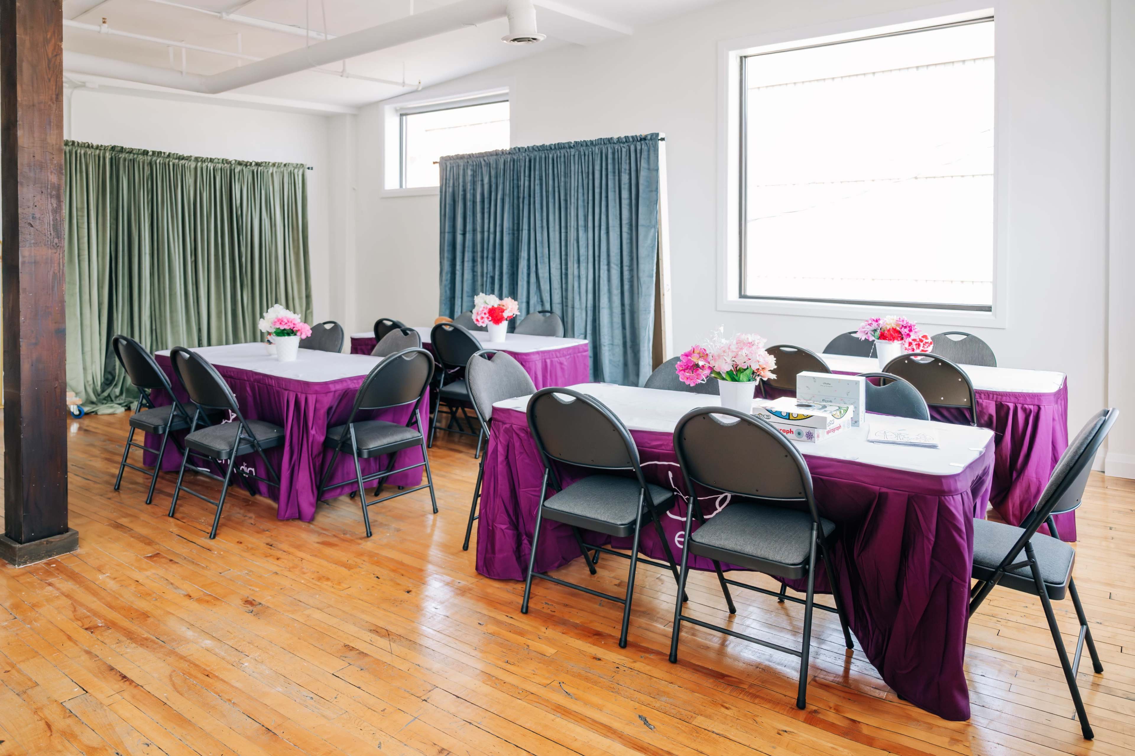 The room features several tables covered with purple cloths and black chairs, surrounded by green and blue curtains, with floral centerpieces on the tables.