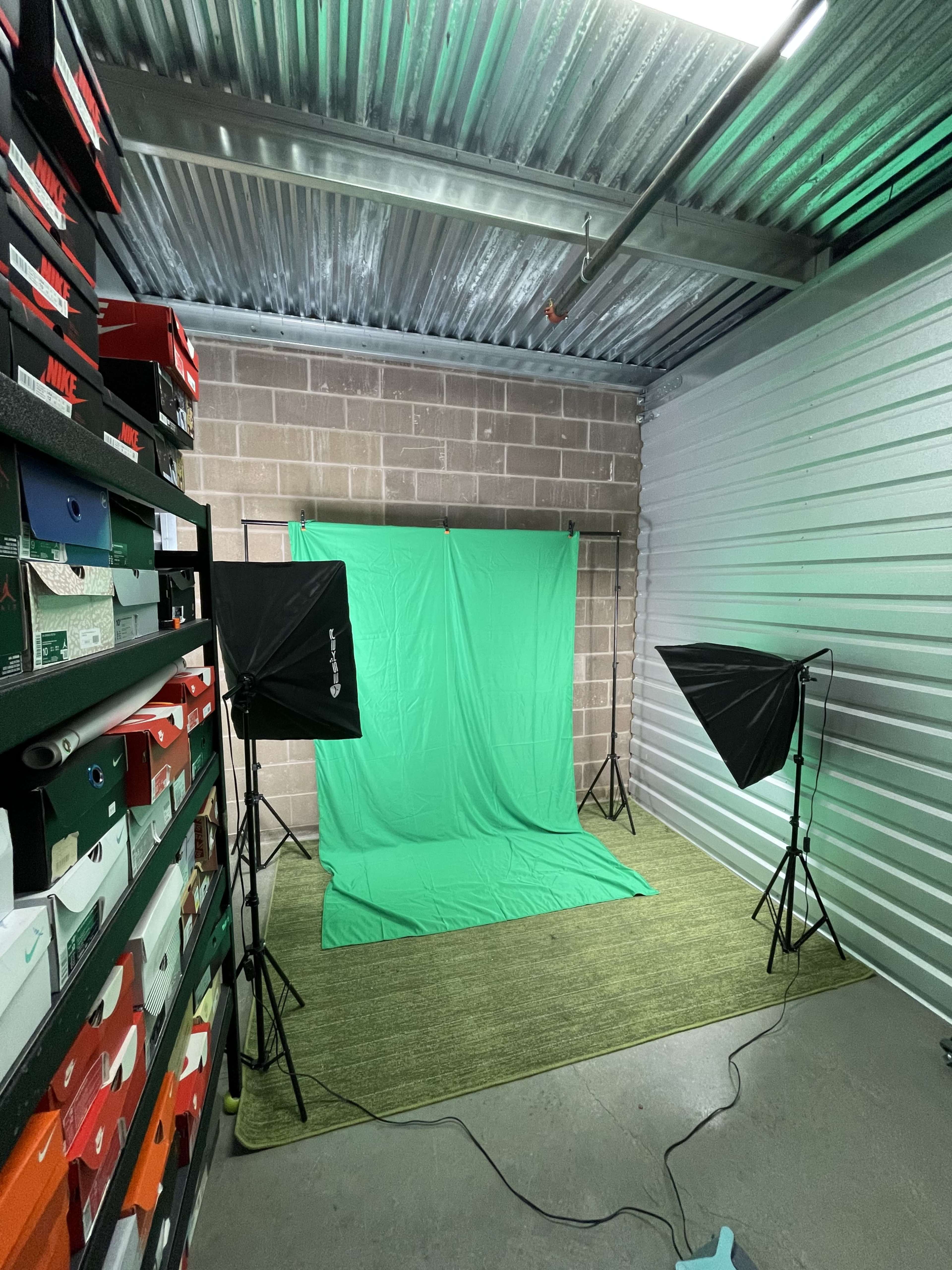 A storage unit contains a green backdrop set up for photography, flanked by two softbox lights and surrounded by stacked shoeboxes.