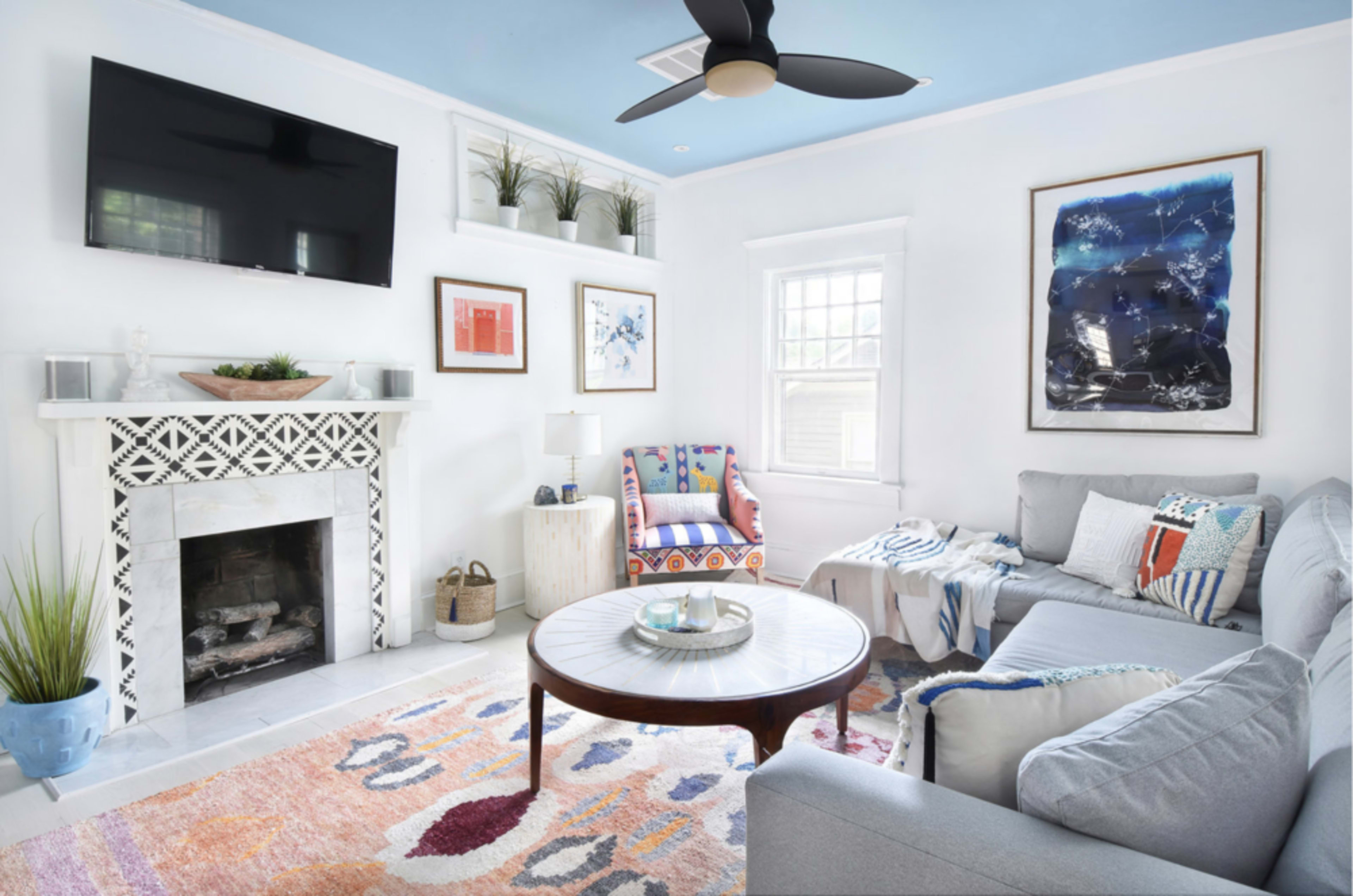 A modern living room features a gray sectional sofa, a round wooden coffee table, a decorative fireplace, and colorful artwork on the walls.