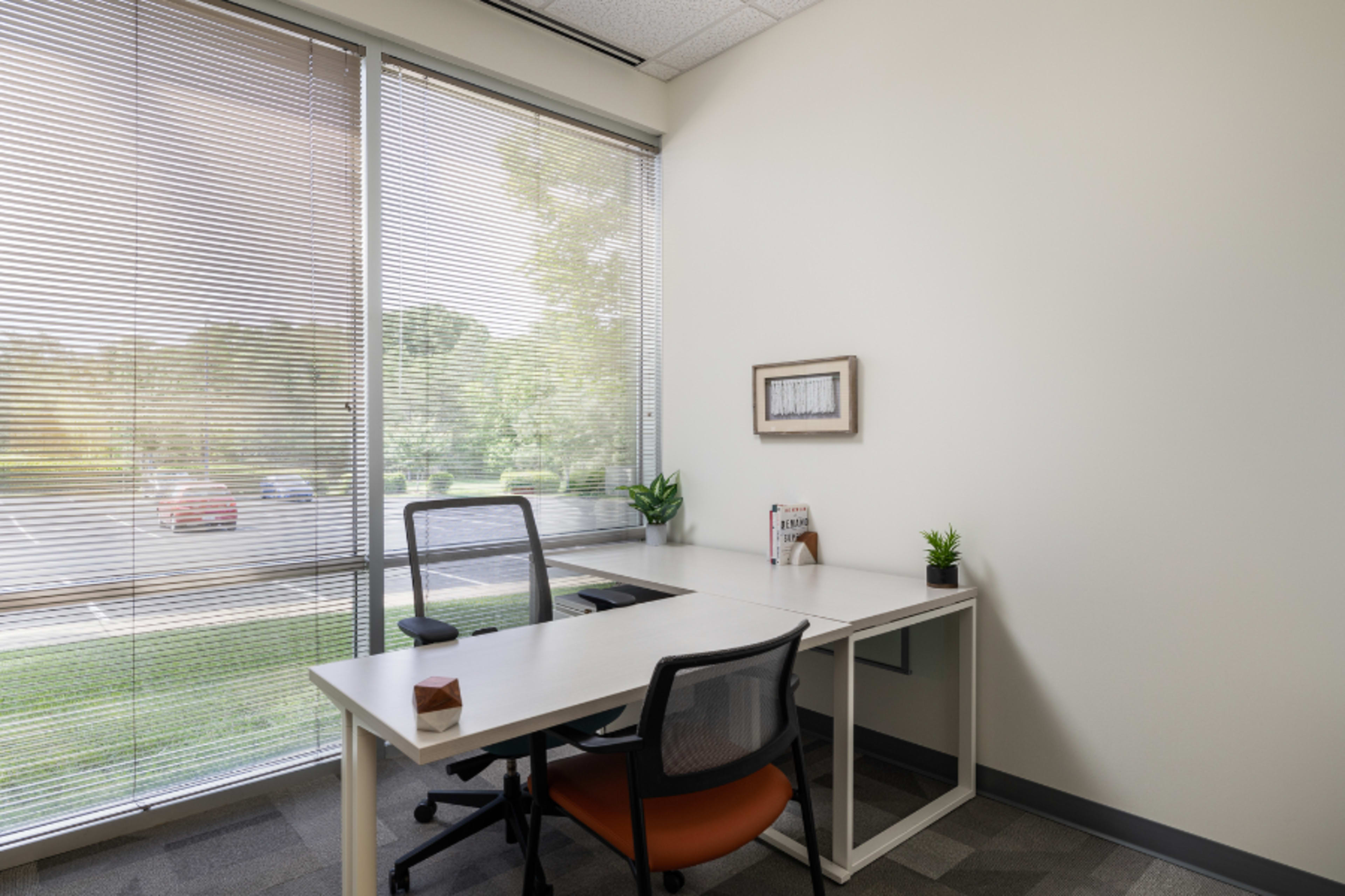 A modern office space with a desk, two chairs, and a large window featuring horizontal blinds.