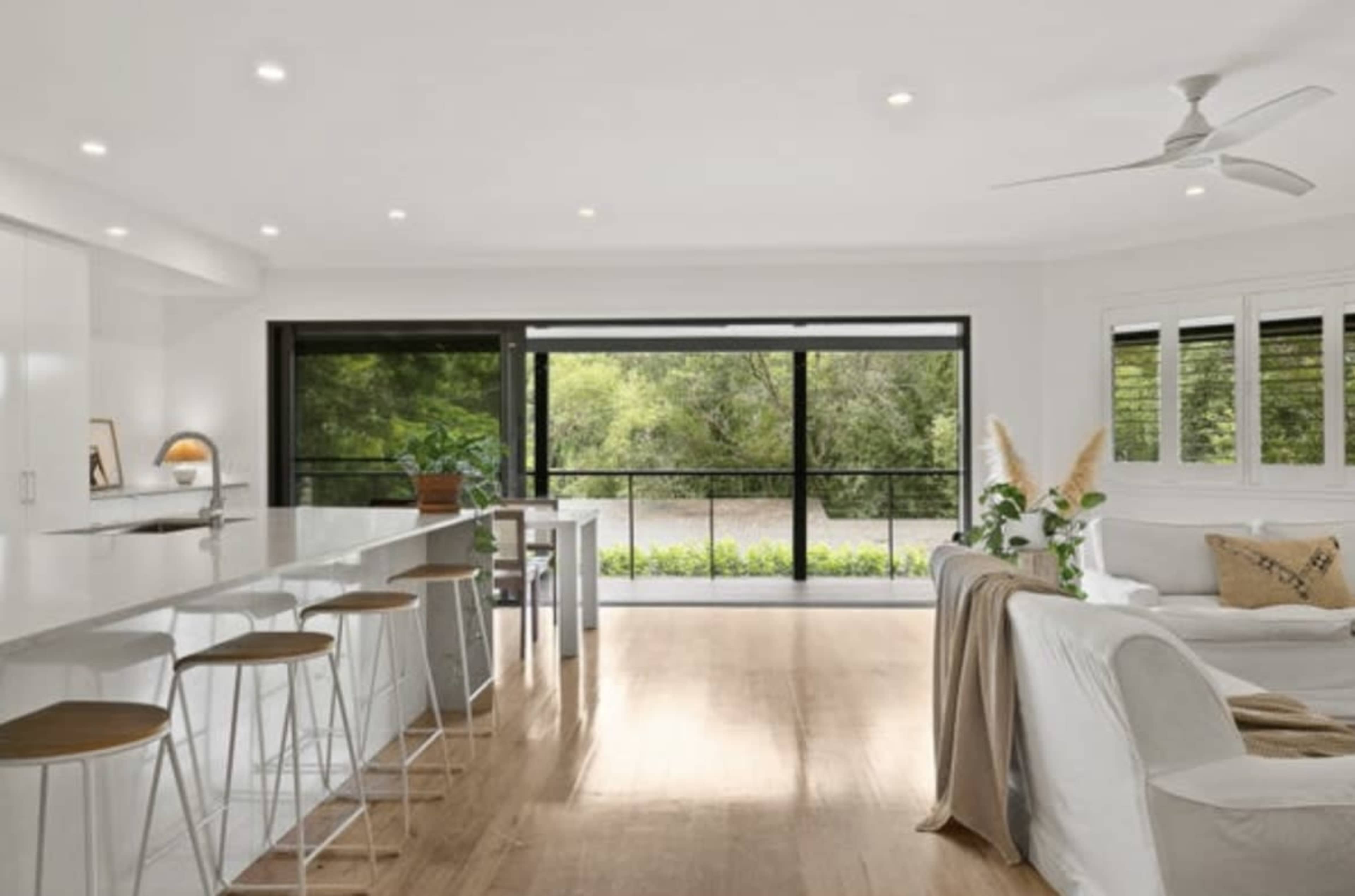 A modern living space features a spacious kitchen with a long island bar, large windows overlooking greenery, and a comfortable seating area.