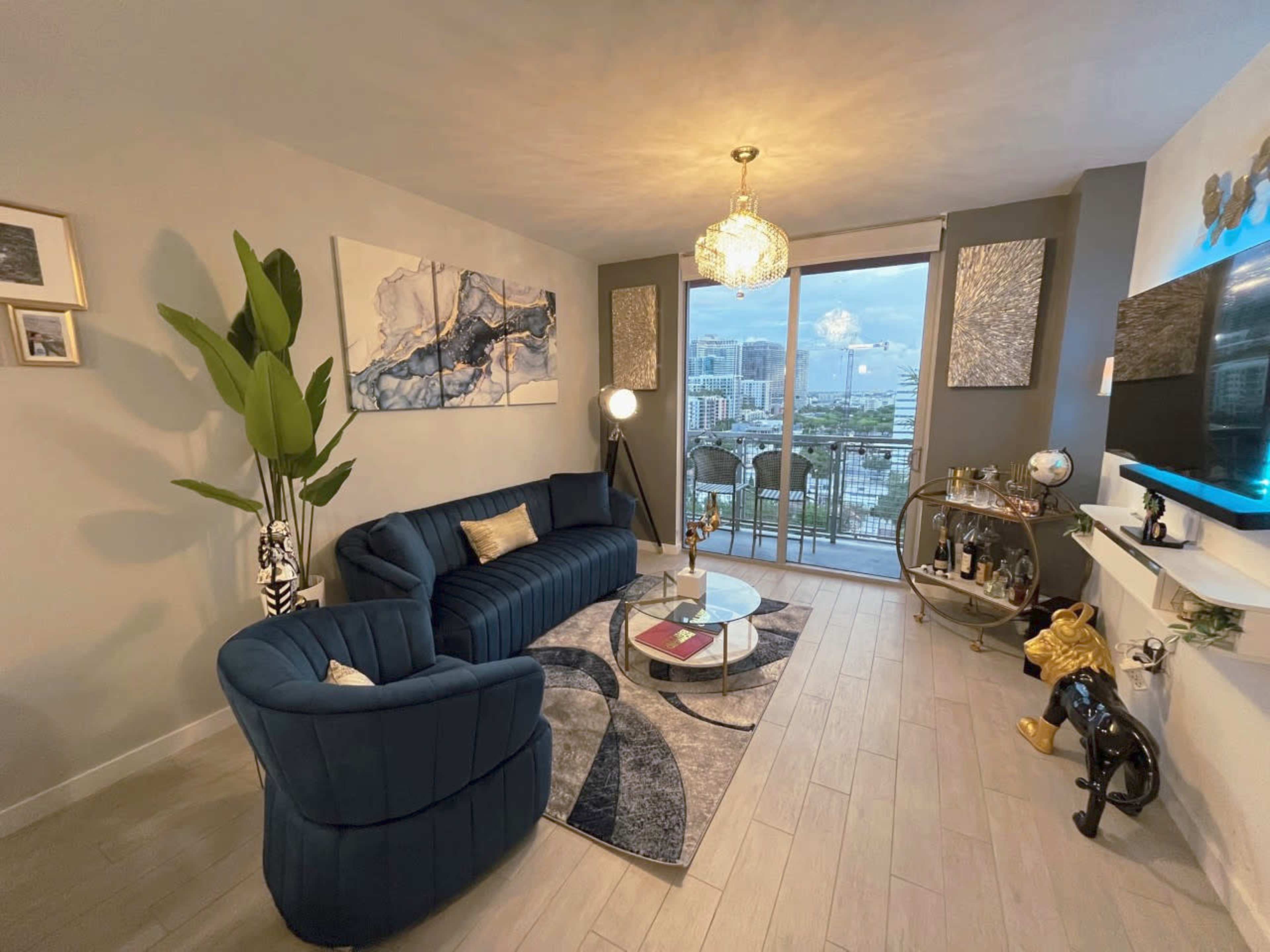 The image shows a modern living room with a blue couch, a glass coffee table, and large windows overlooking a cityscape.