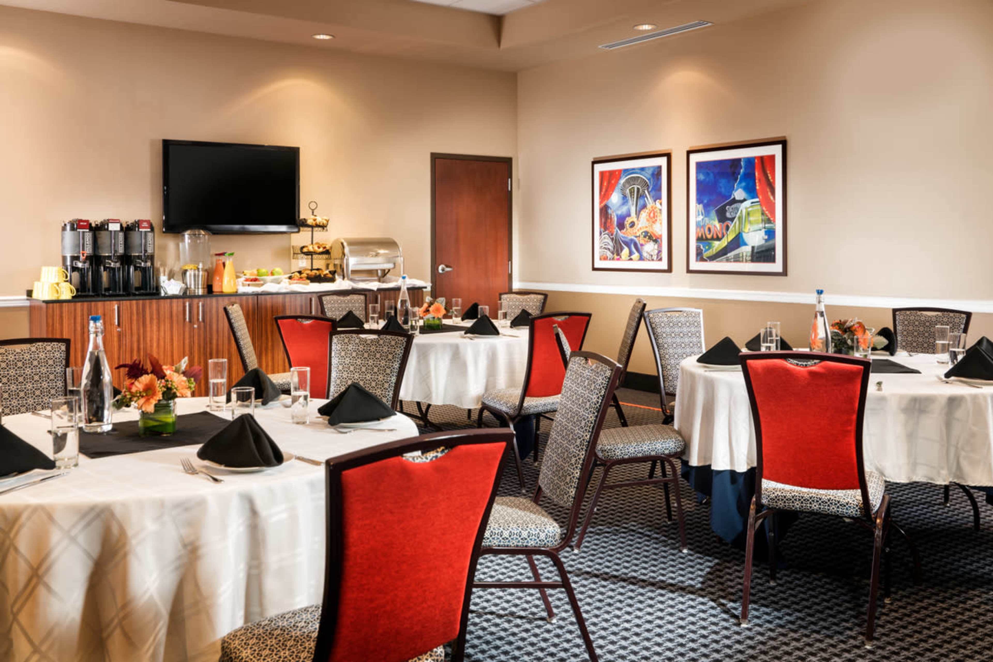 A dining room setup with round tables covered in white tablecloths, each adorned with black napkins, surrounded by chairs and featuring a buffet table in the background.