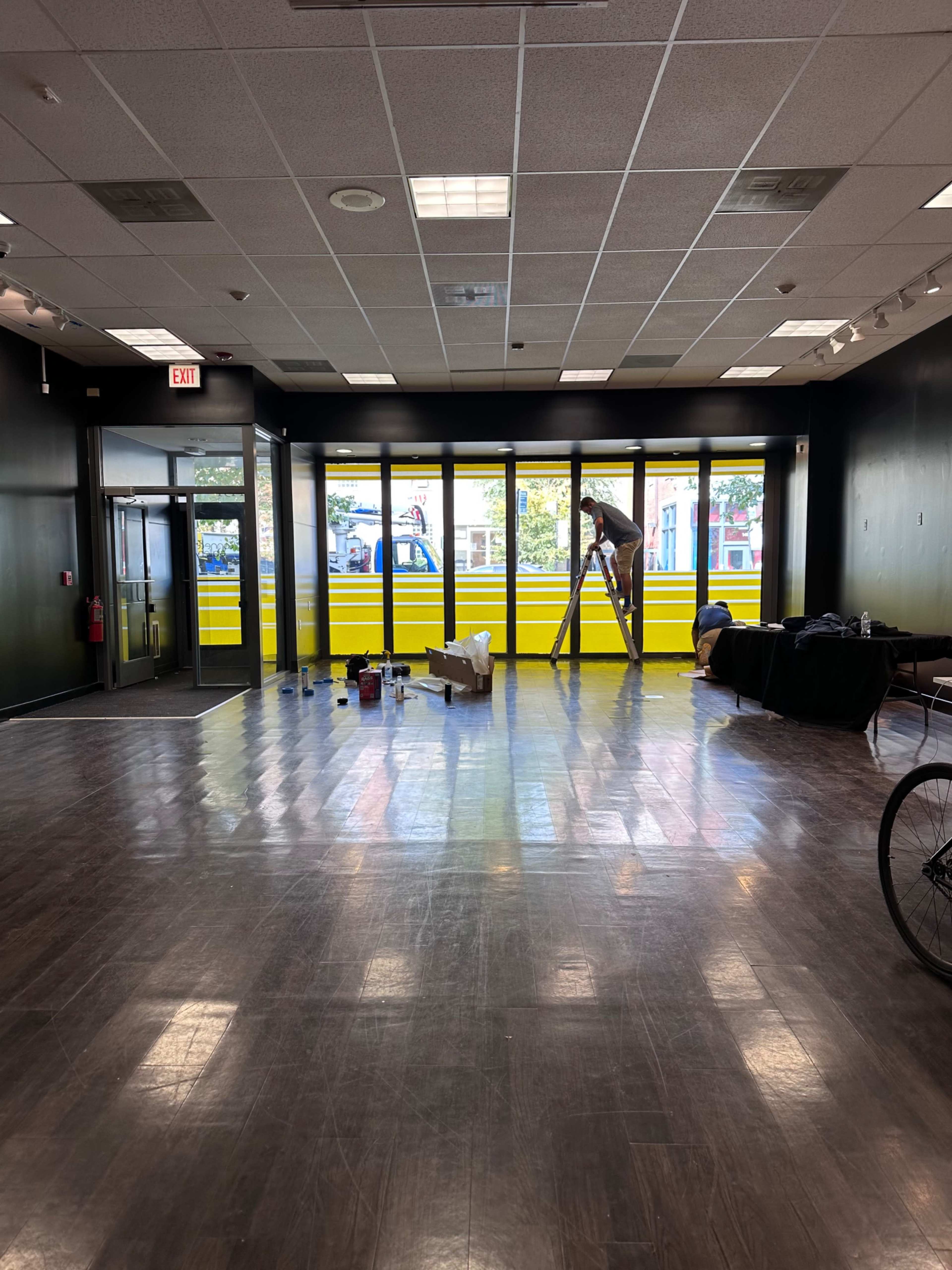 The image shows a spacious room with a wooden floor, featuring a person on a ladder painting yellow panels on the large front windows, while various items are scattered on the floor.