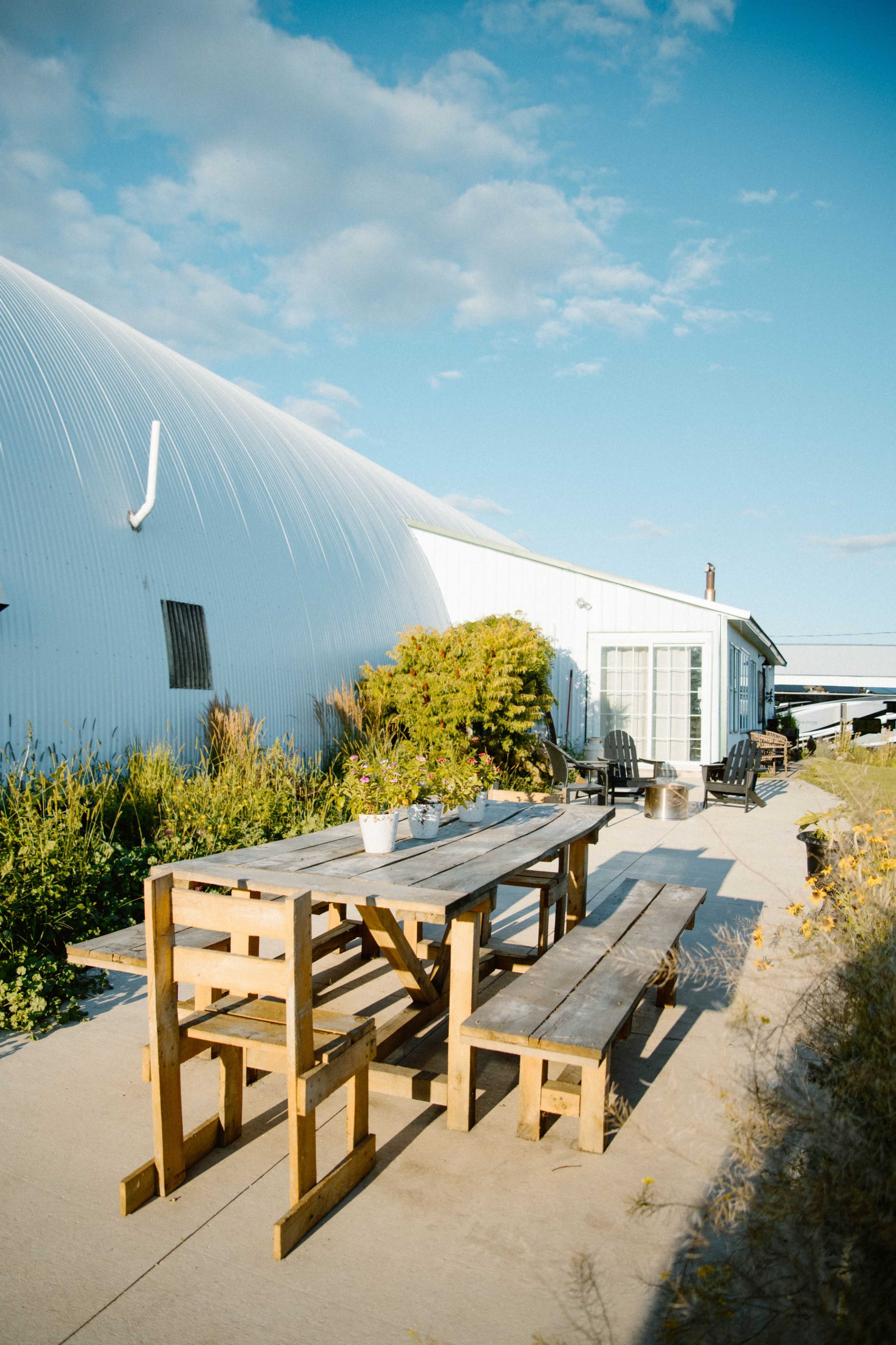 A wooden table and benches sit outside a white, curved building, surrounded by greenery and under a clear blue sky.
