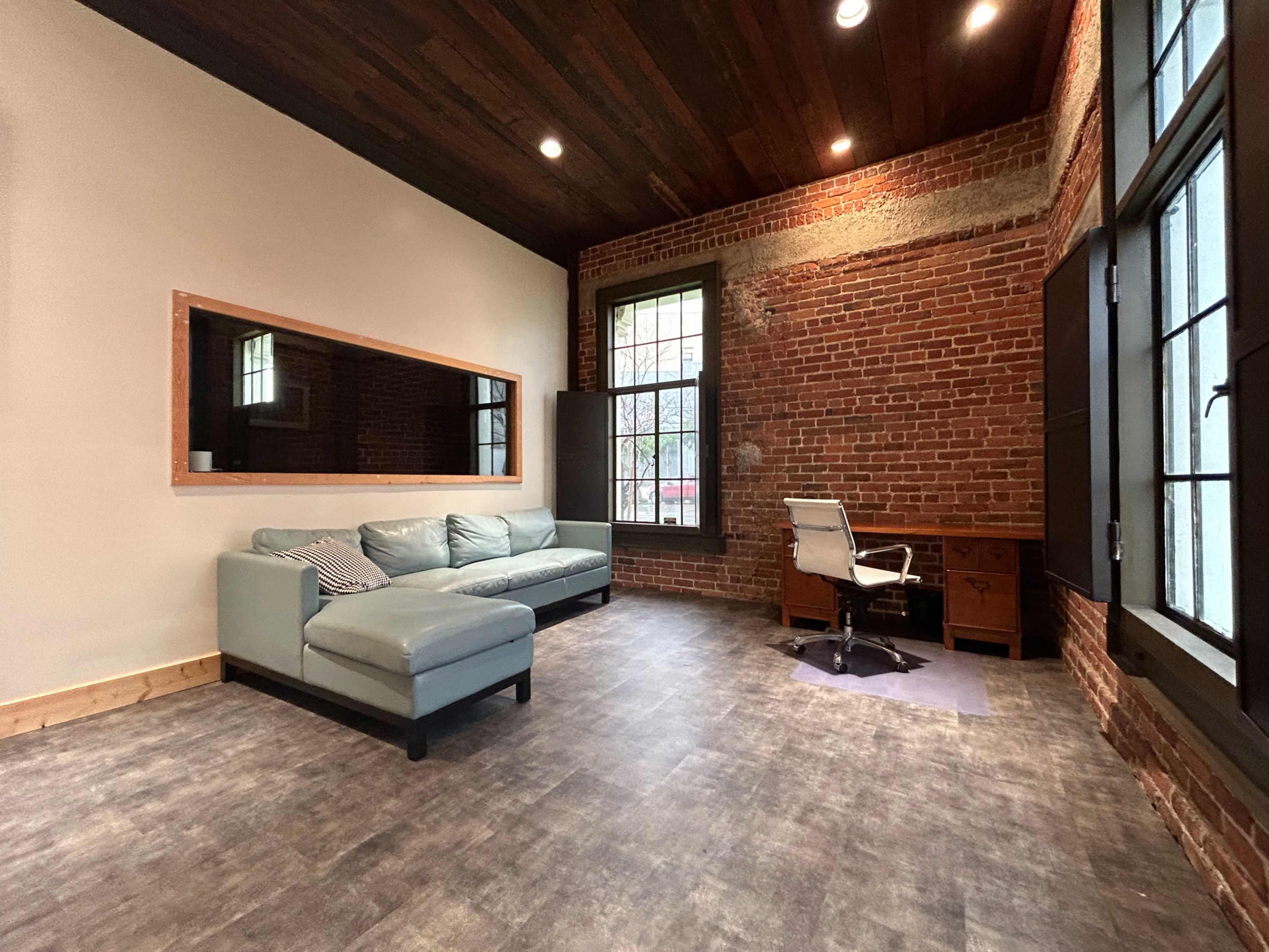 A modern workspace featuring a light blue couch, a desk with a chair, and exposed brick walls with wooden beams on the ceiling.