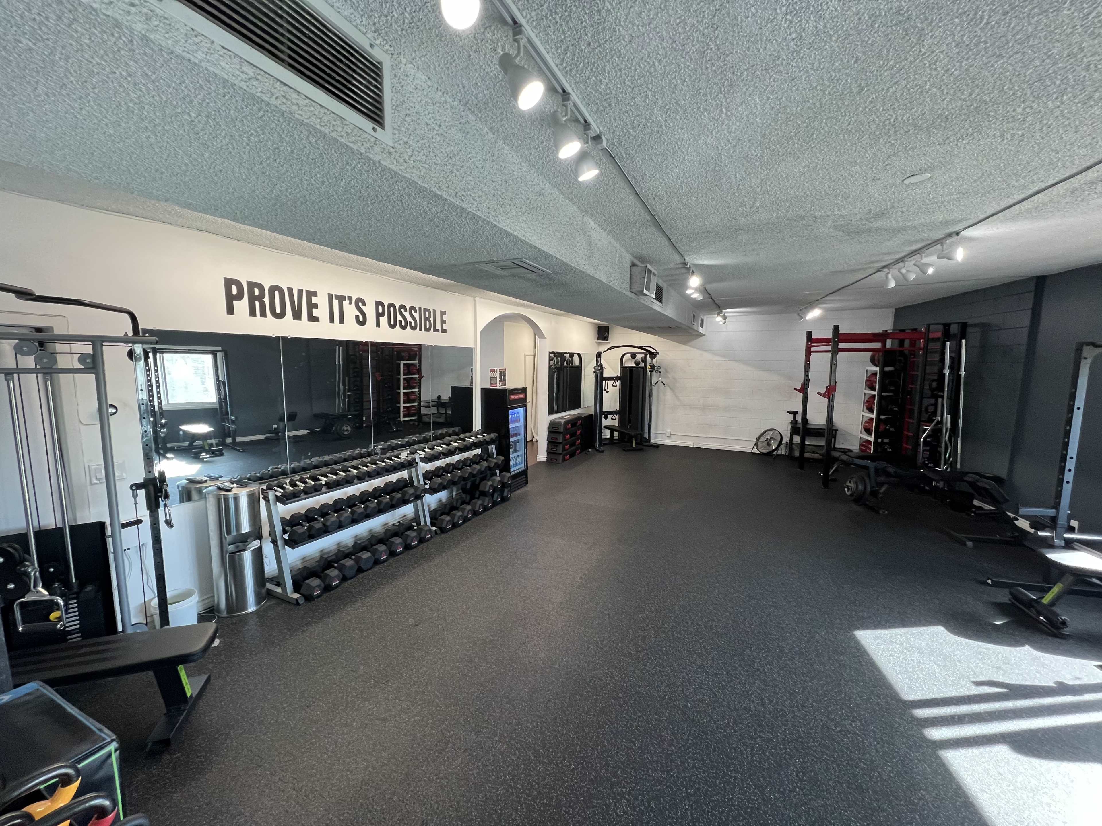 The image shows a well-equipped gym interior featuring weight lifting machines, dumbbells, and a motivational phrase on the wall.
