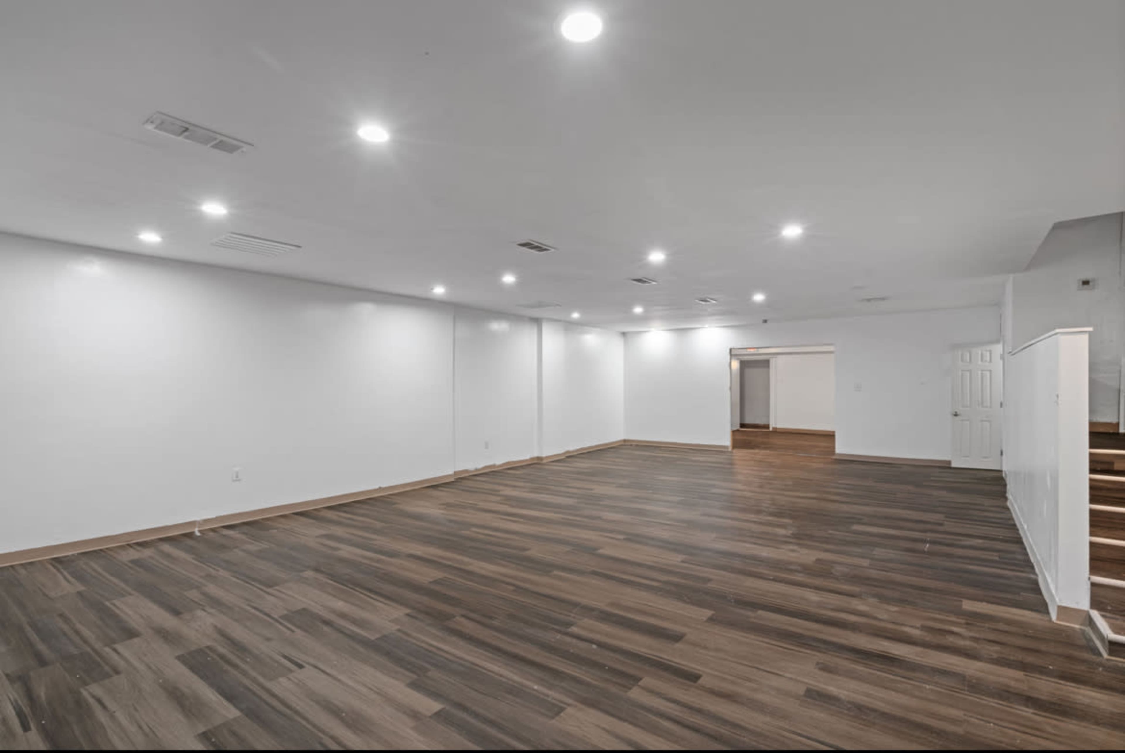 The image shows a spacious, empty room with wooden flooring and bright overhead lighting.