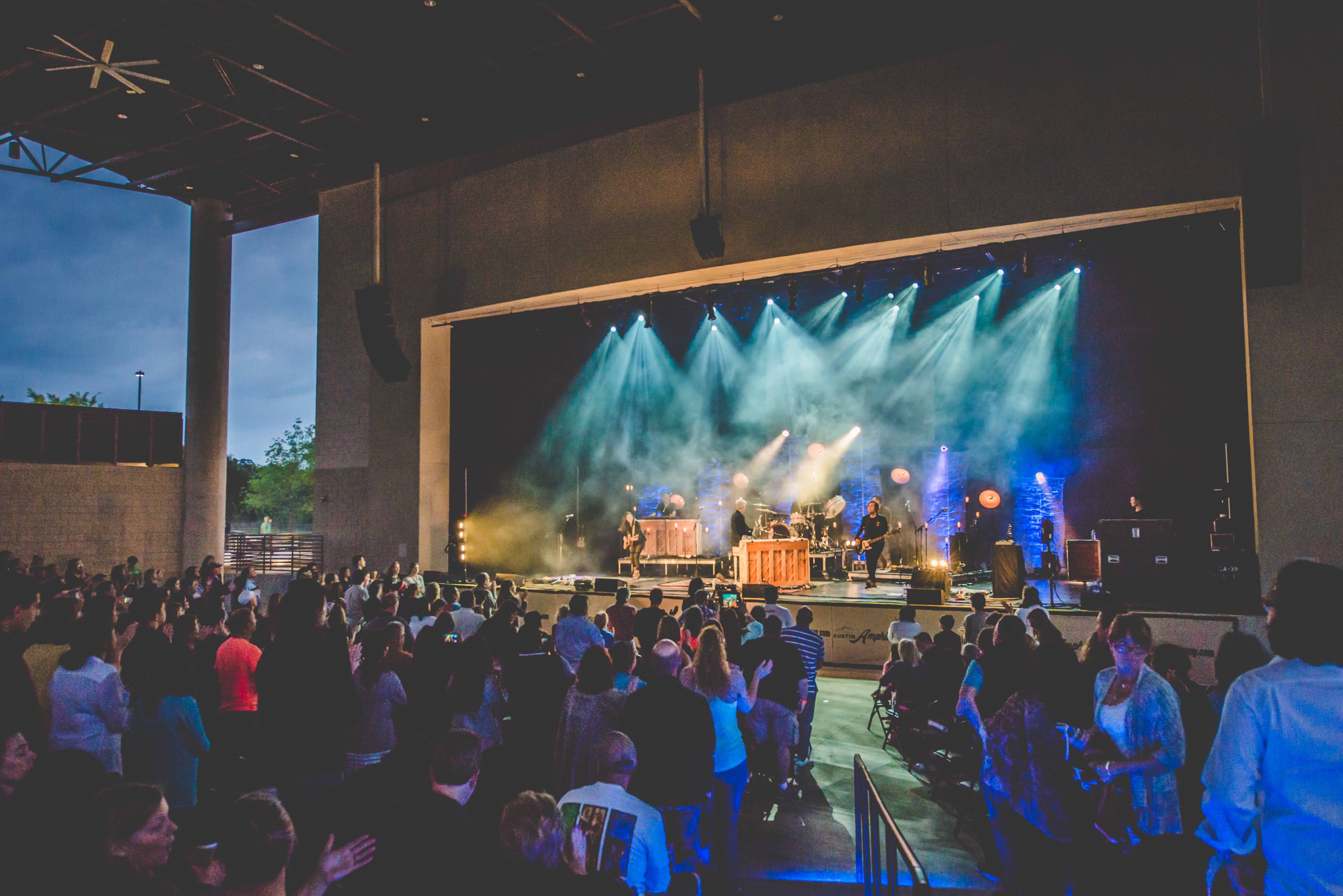 A live music concert is taking place on an outdoor stage with a crowd watching and colorful lights illuminating the performers.