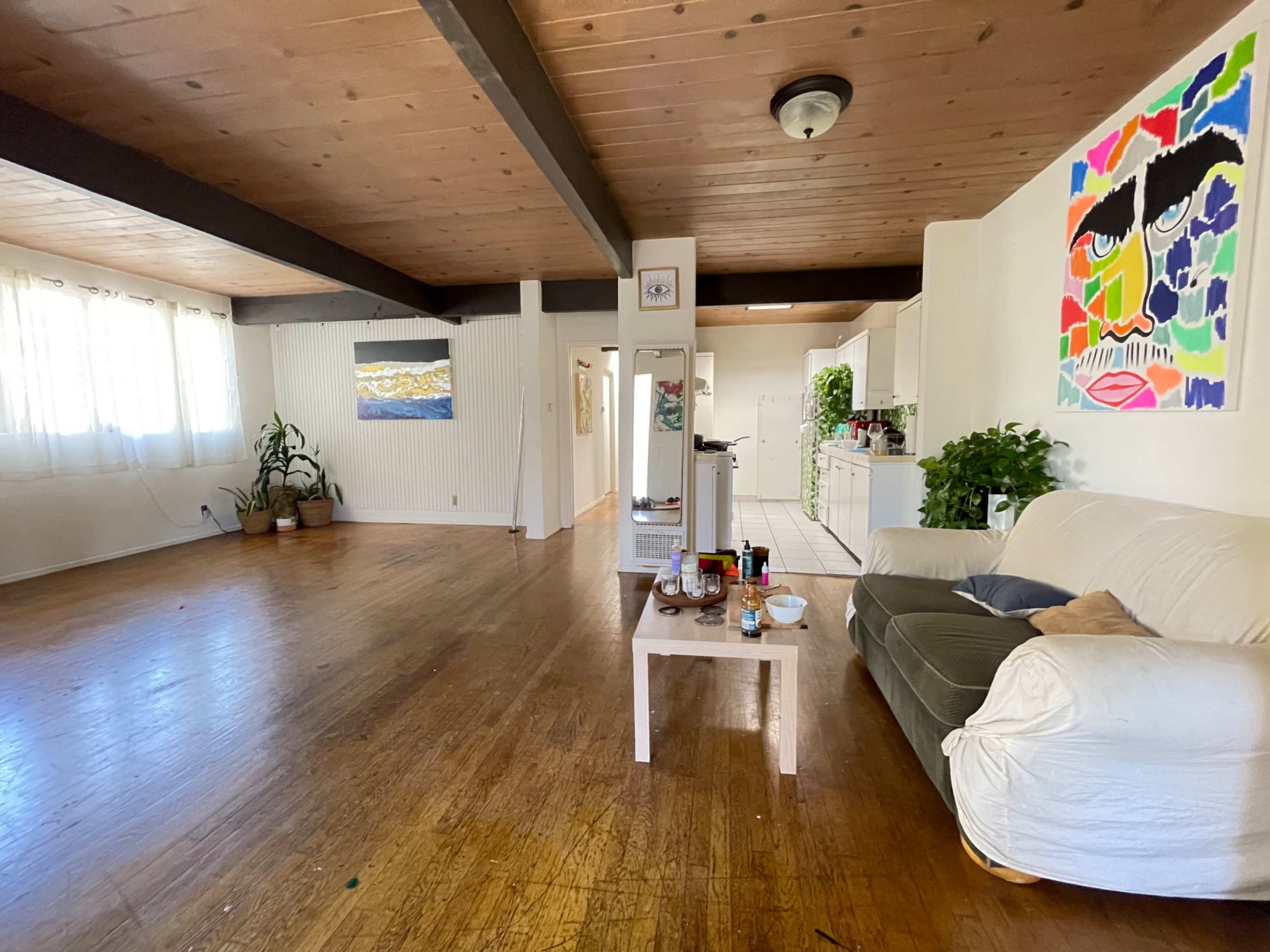 A spacious living room with wooden floors, a white couch, a coffee table with items on it, and large windows allowing natural light to enter.