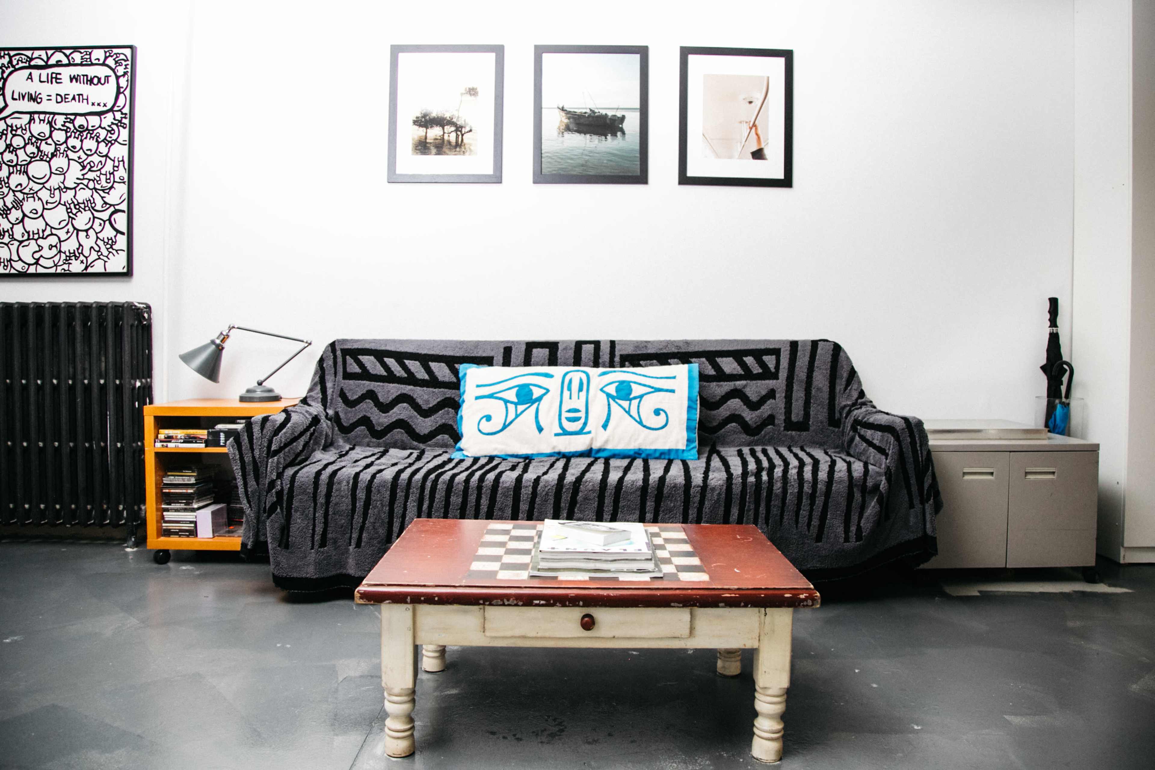 The image shows a living room with a black and white patterned couch, a coffee table with a magazine, a lamp, and several framed pictures on the wall.