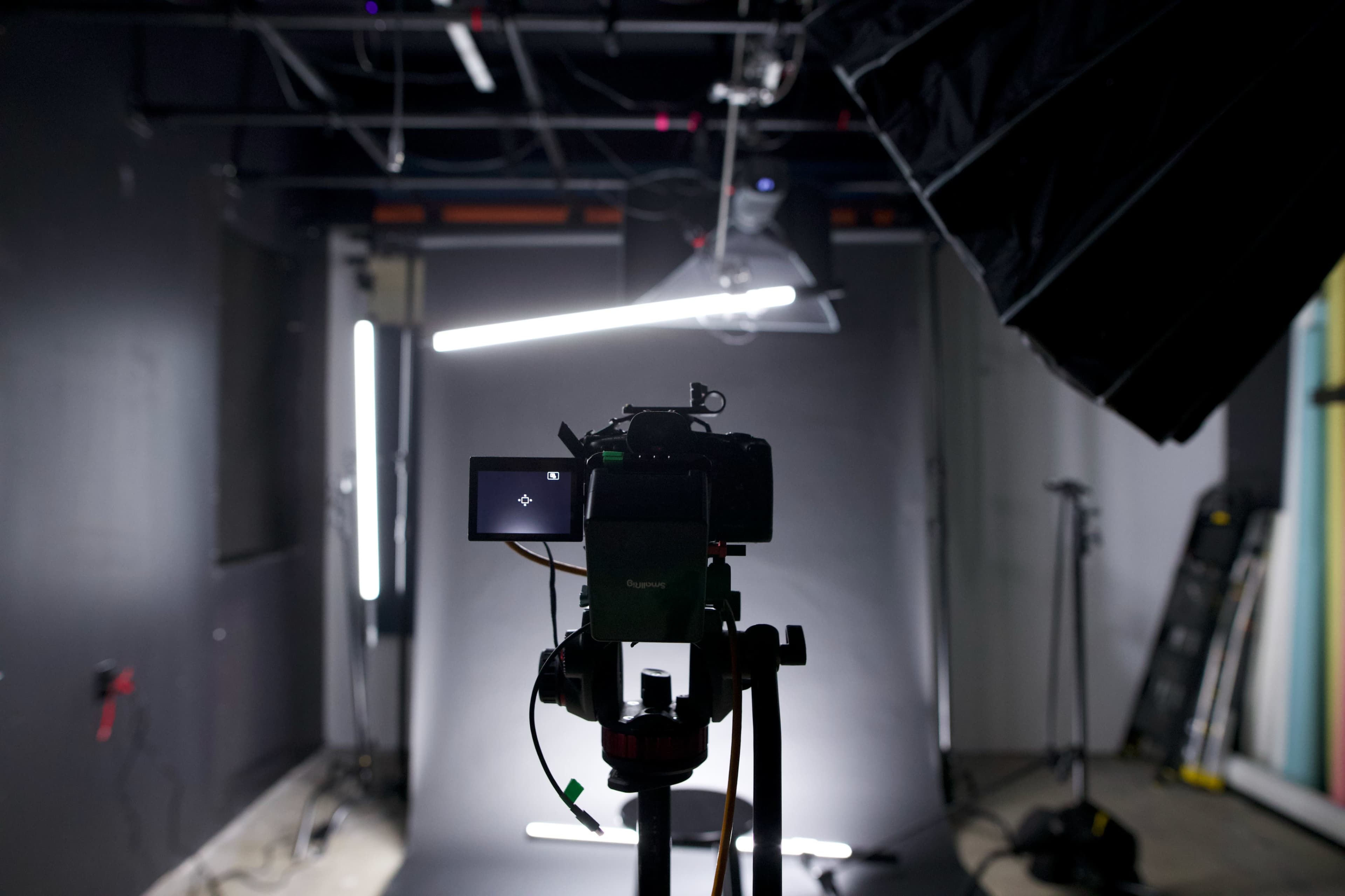A camera is positioned on a tripod in a dimly lit studio, facing a backdrop with lighting equipment visible.