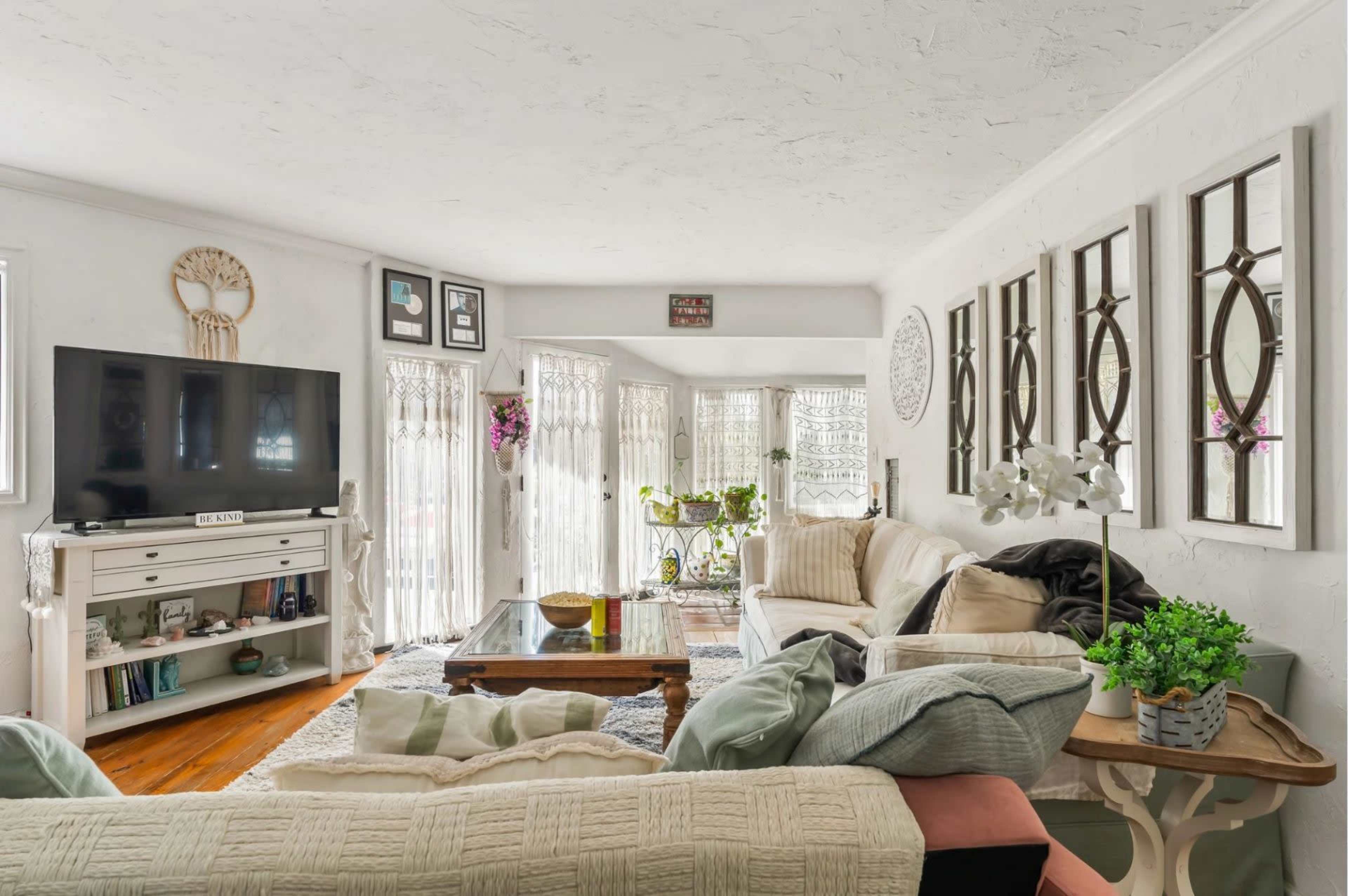 The living room features a large television, a sectional sofa with various cushions, a coffee table, and windows adorned with lace curtains letting in natural light.