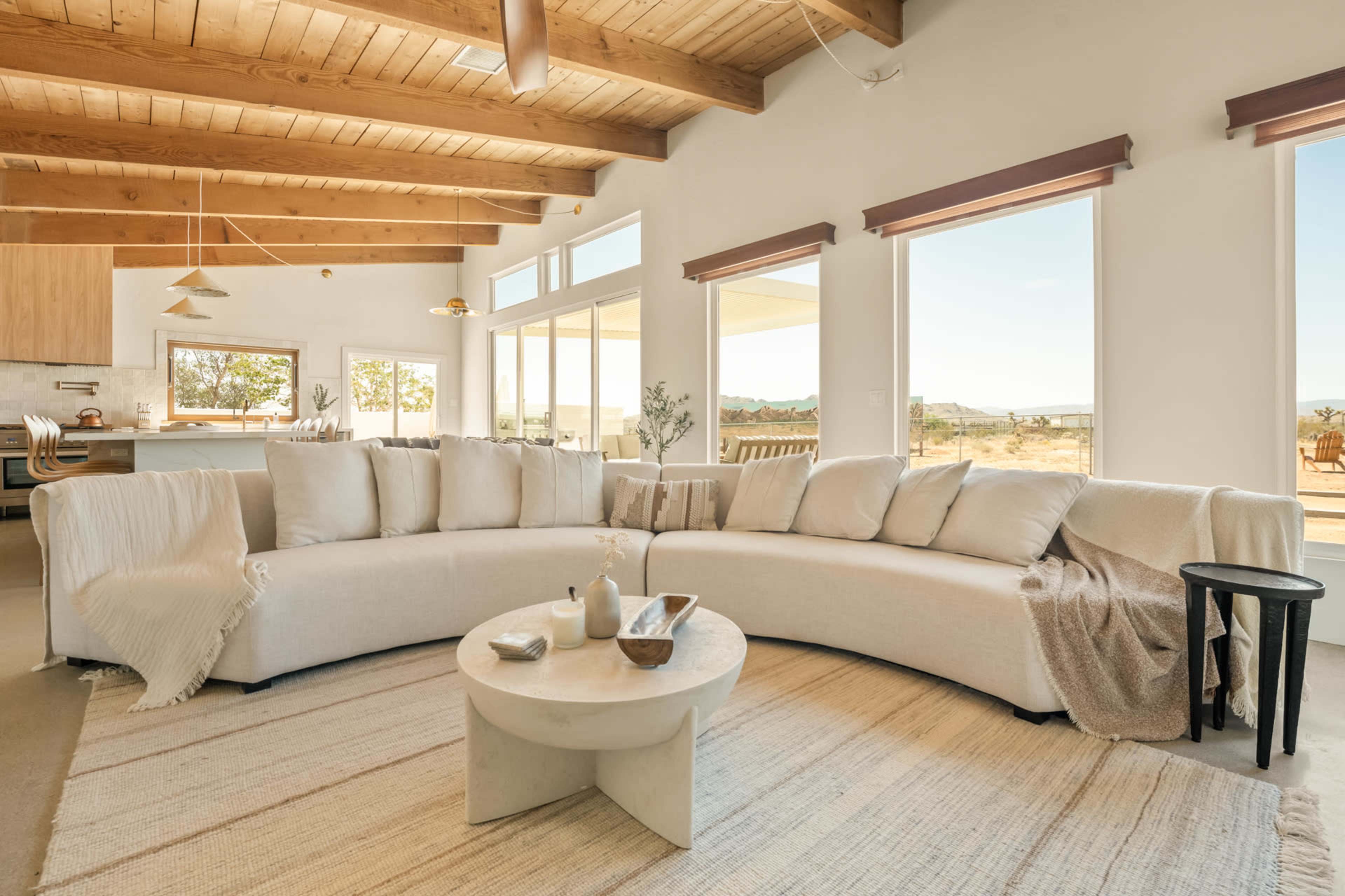 A spacious living area features a curved beige couch, a round coffee table, and large windows providing views of the outdoors.