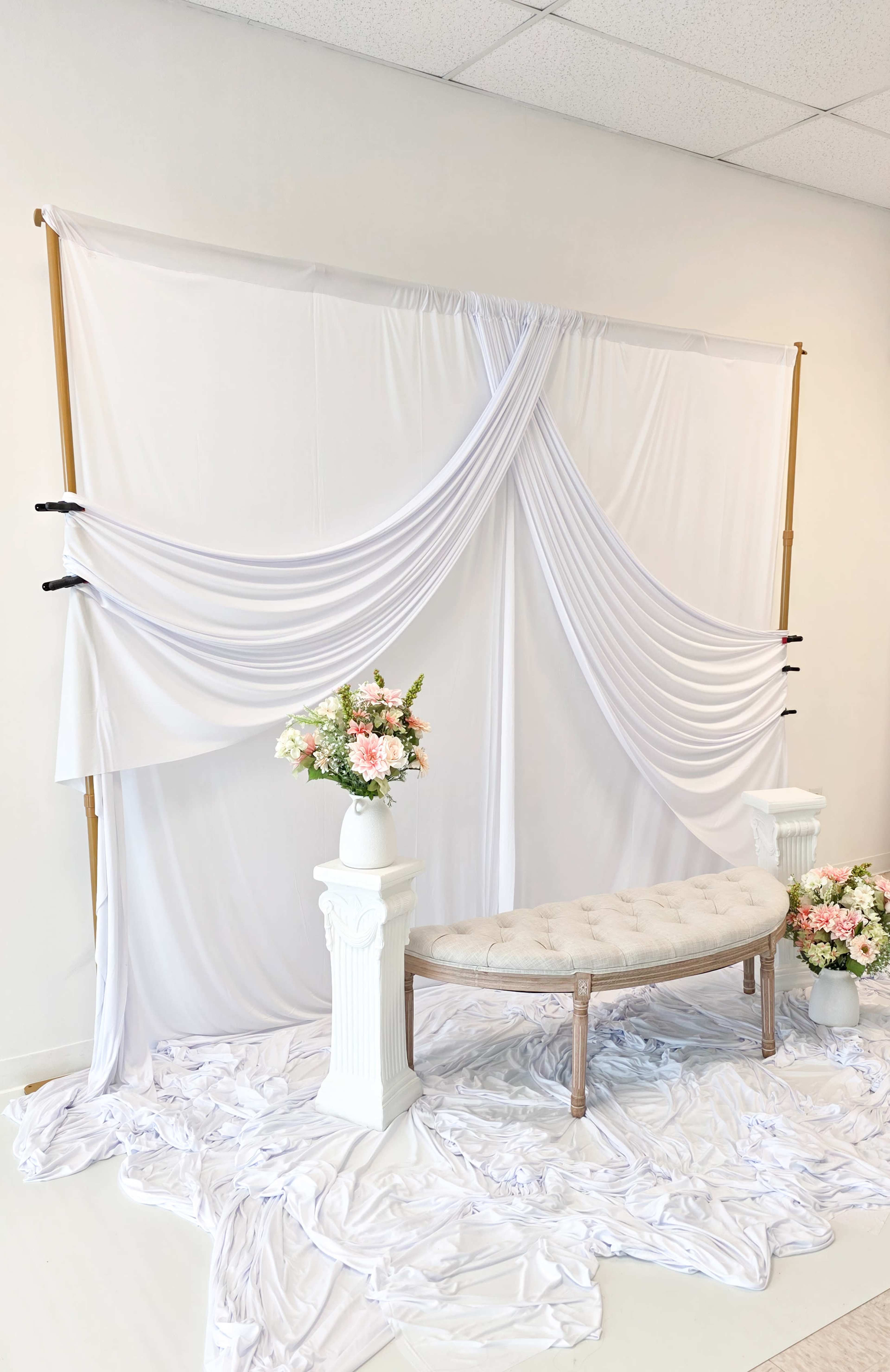 The image features a white draped backdrop with a decorative bench and floral arrangements on ornate pedestals.