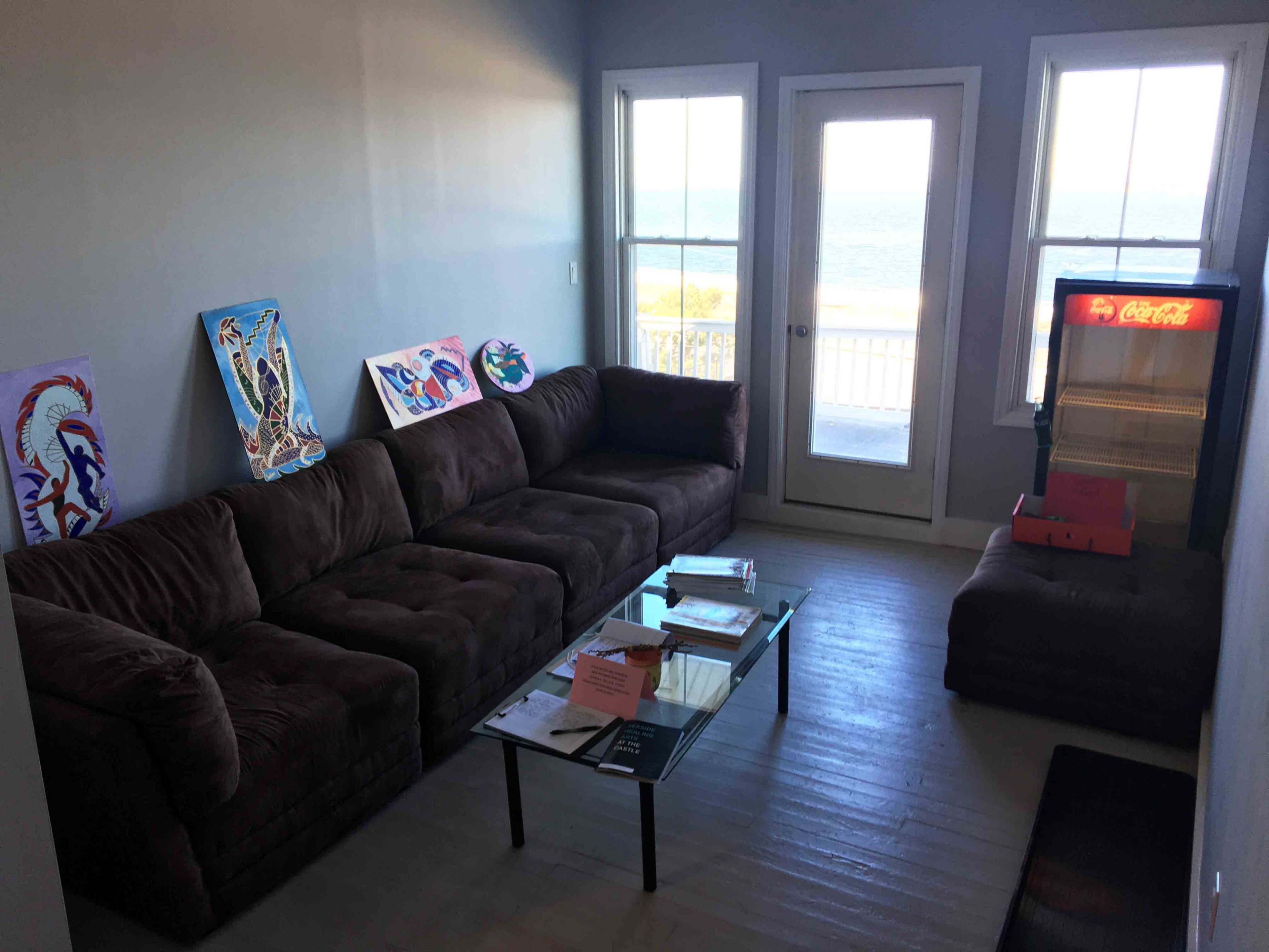 A living room features a brown couch, a glass coffee table with papers, and a Coca-Cola refrigerator, with large windows showing a view of the ocean.
