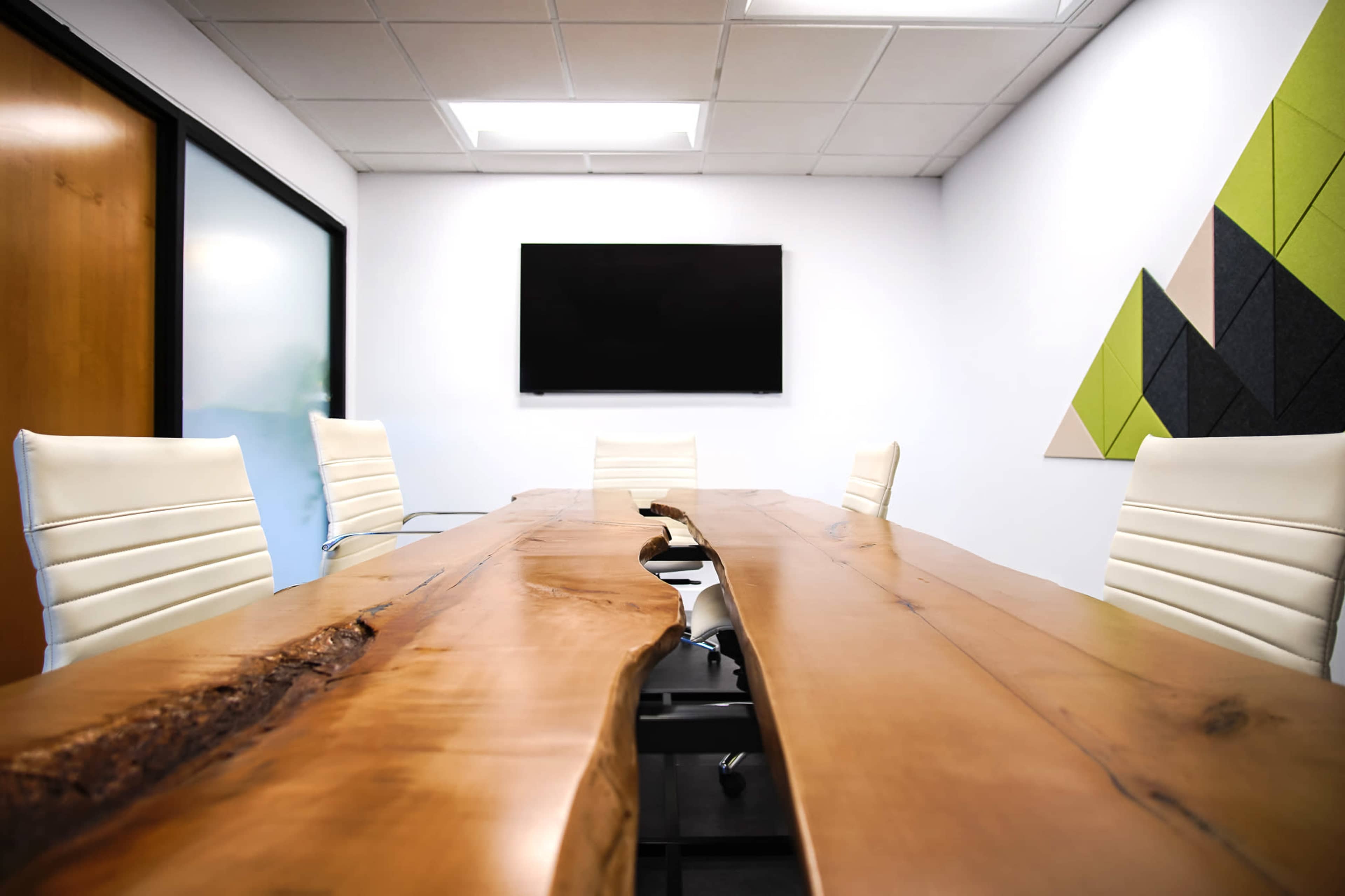 A modern conference room features a long wooden table with a live edge, surrounded by white chairs, and a wall-mounted TV against a backdrop of geometric wall panels.