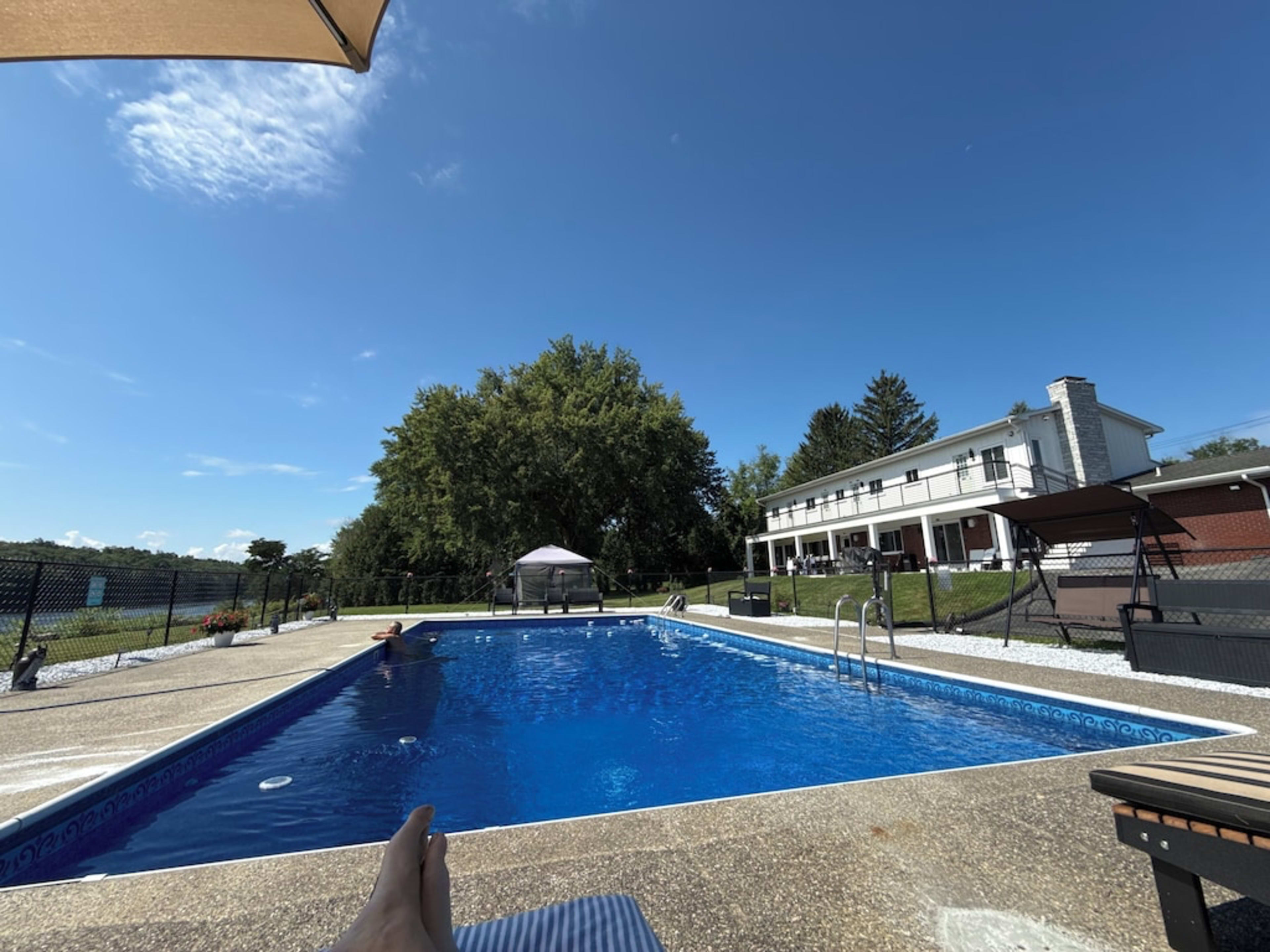The image shows a clear swimming pool surrounded by a deck, with a house and trees in the background under a blue sky.
