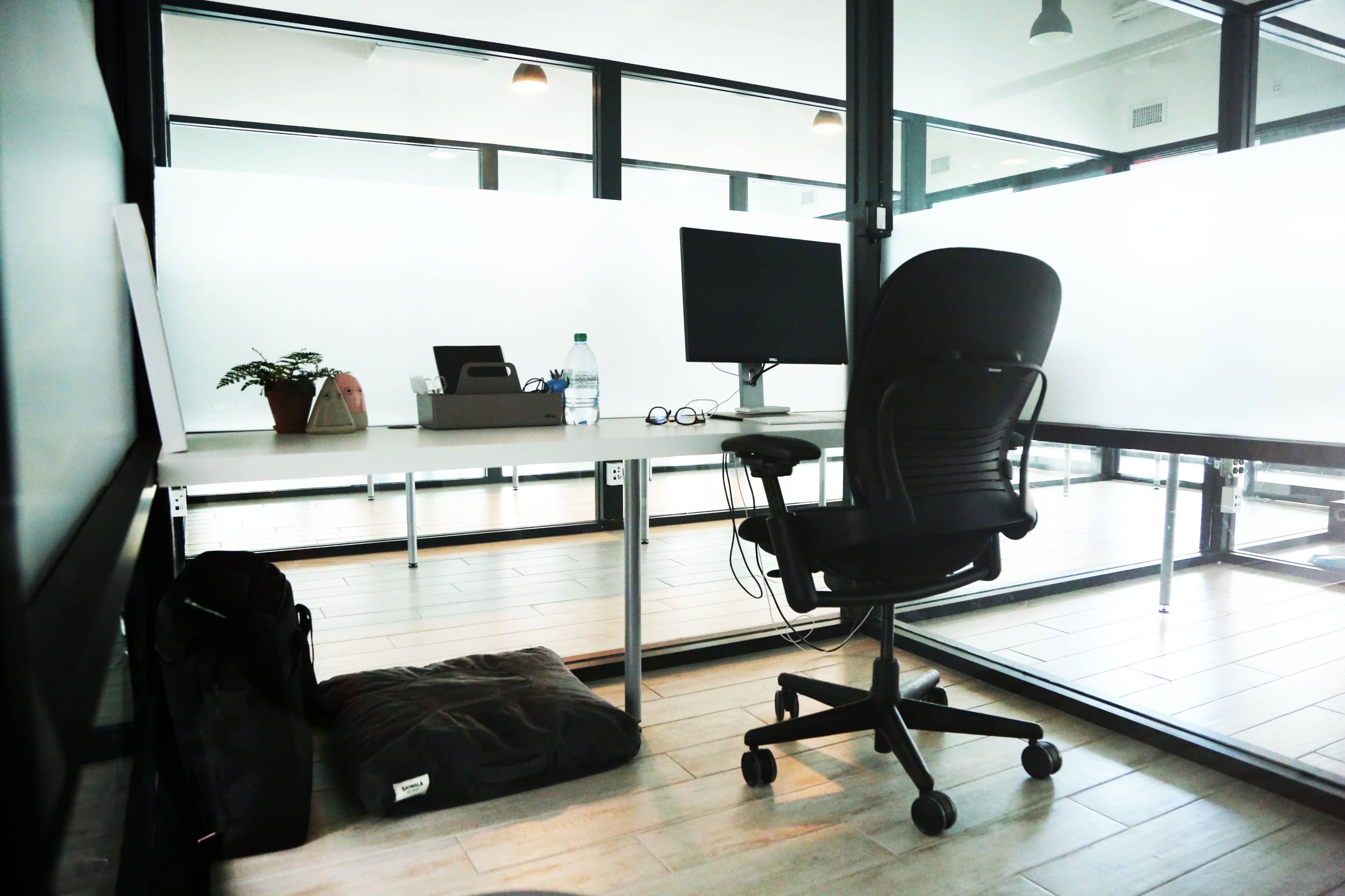 An empty office space features a desk with a computer monitor, a chair, and a small plant, surrounded by glass walls.