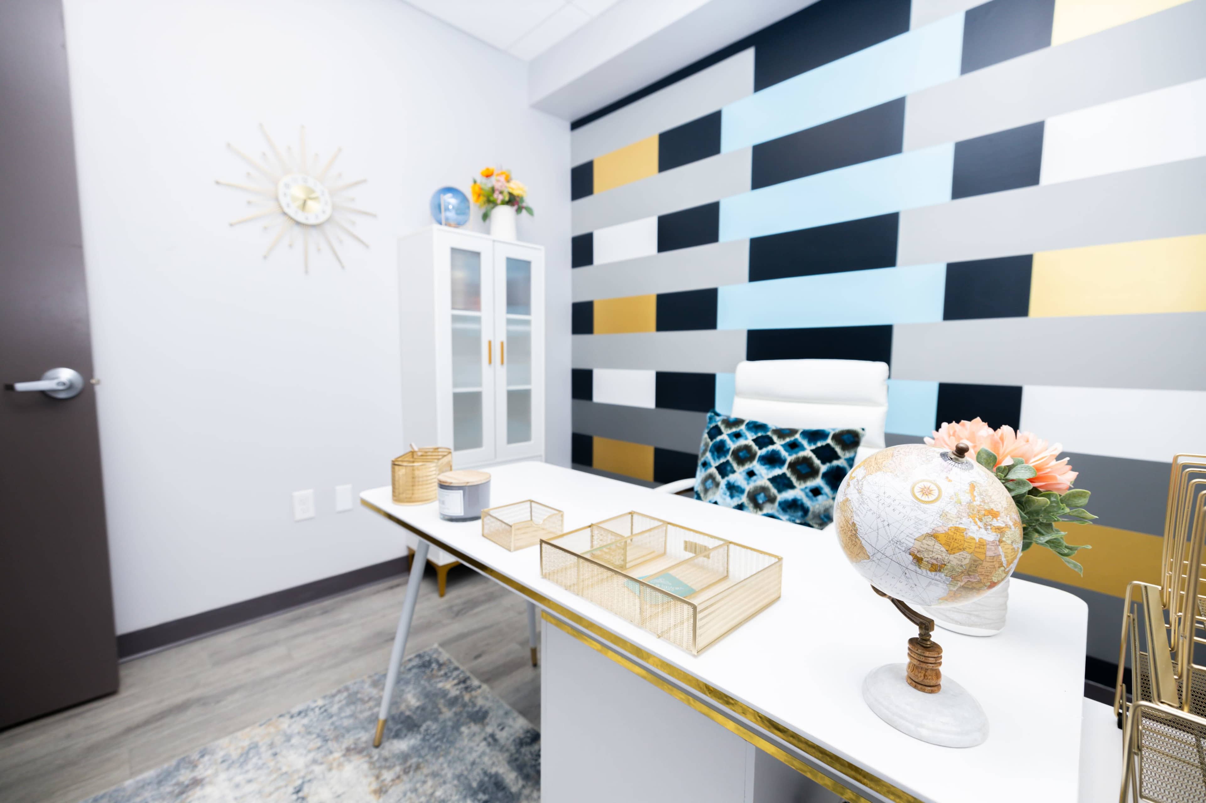 The image shows a modern office with a white desk, decorative globe, and a colorful accent wall featuring geometric patterns.