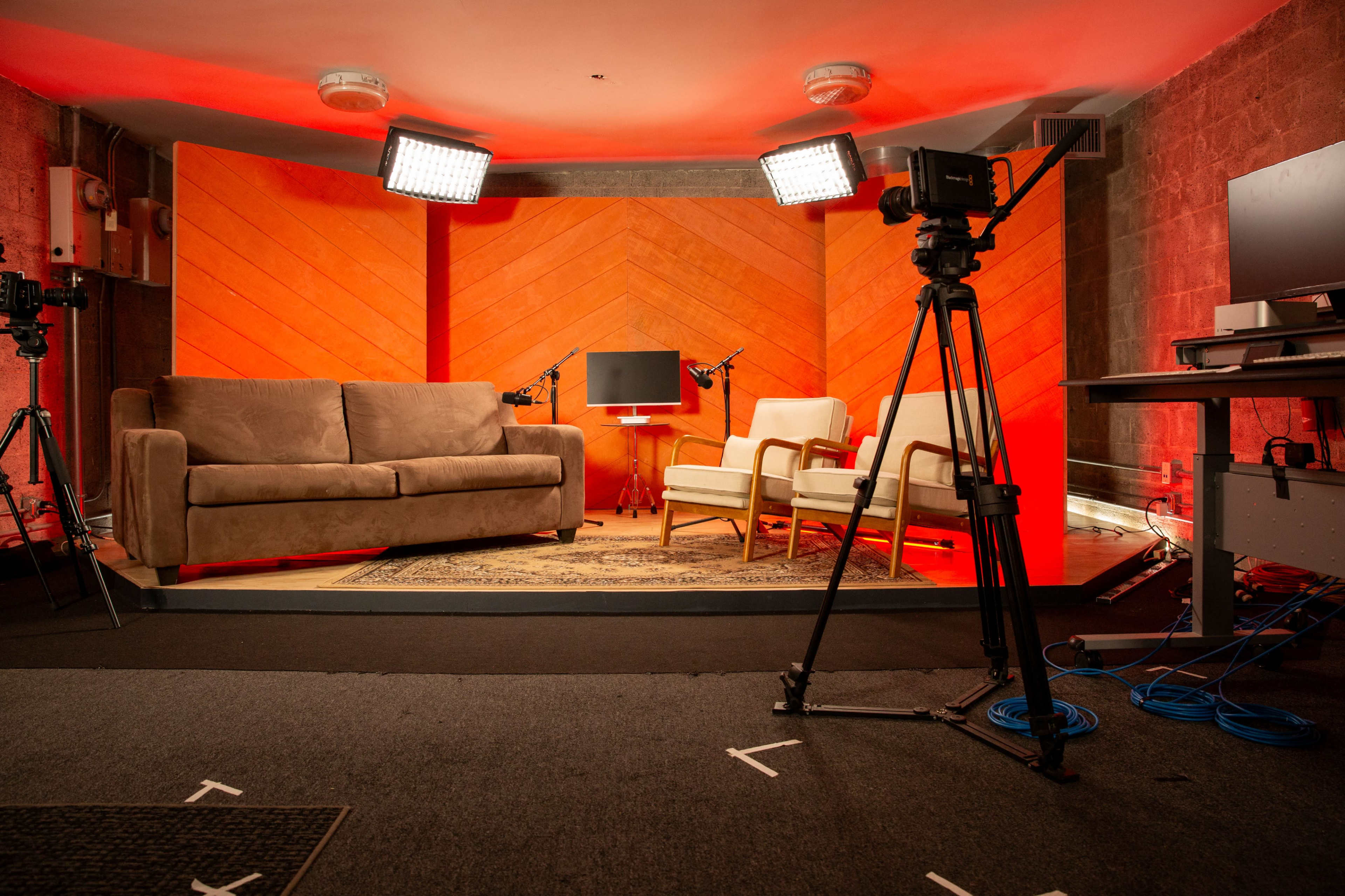 The image shows a studio setup with a sofa, two armchairs, a coffee table, and lighting equipment against an orange backdrop.