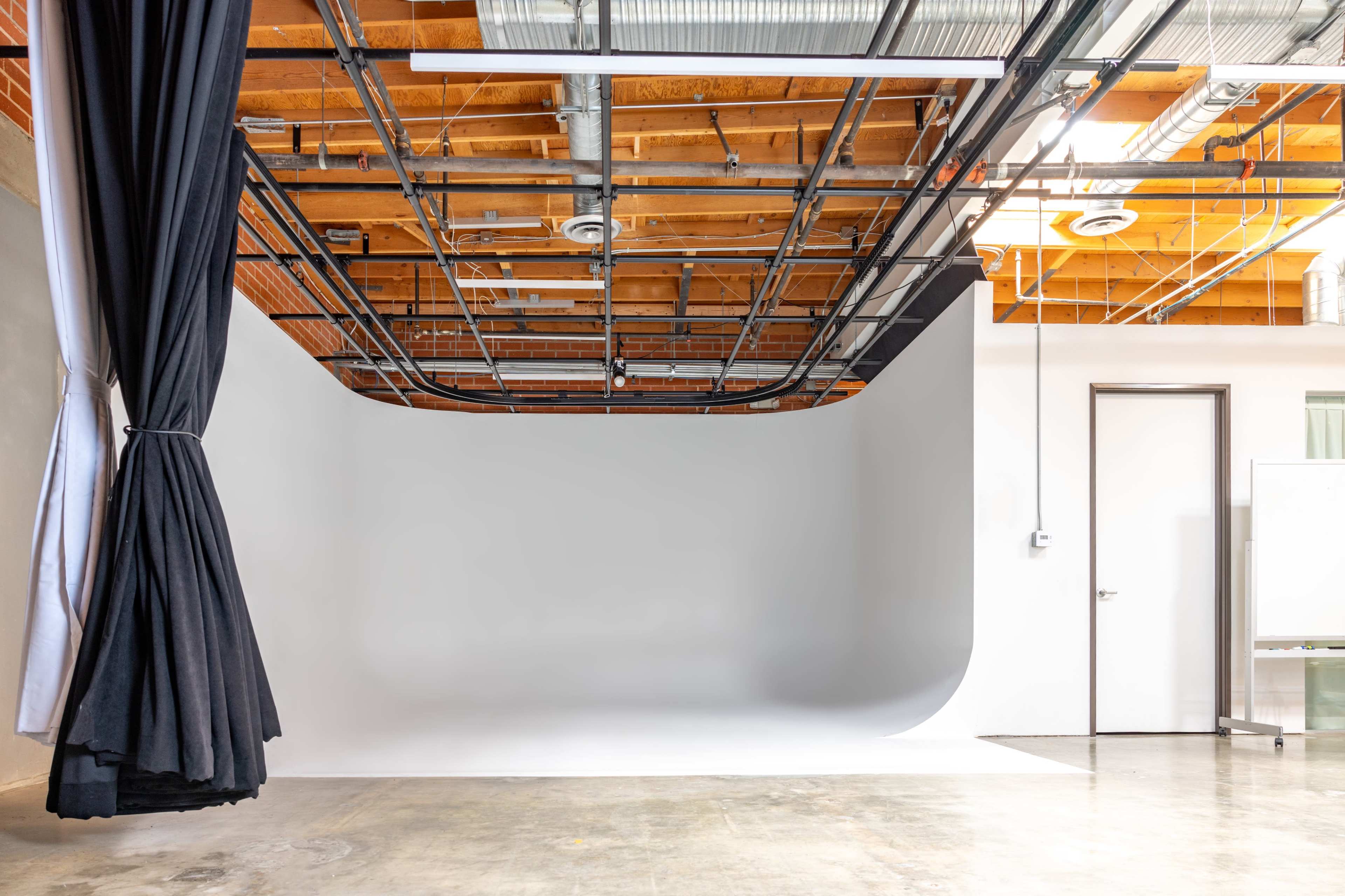 The image shows a photography studio with a seamless white backdrop and black curtains, featuring exposed wooden beams and metal piping overhead.