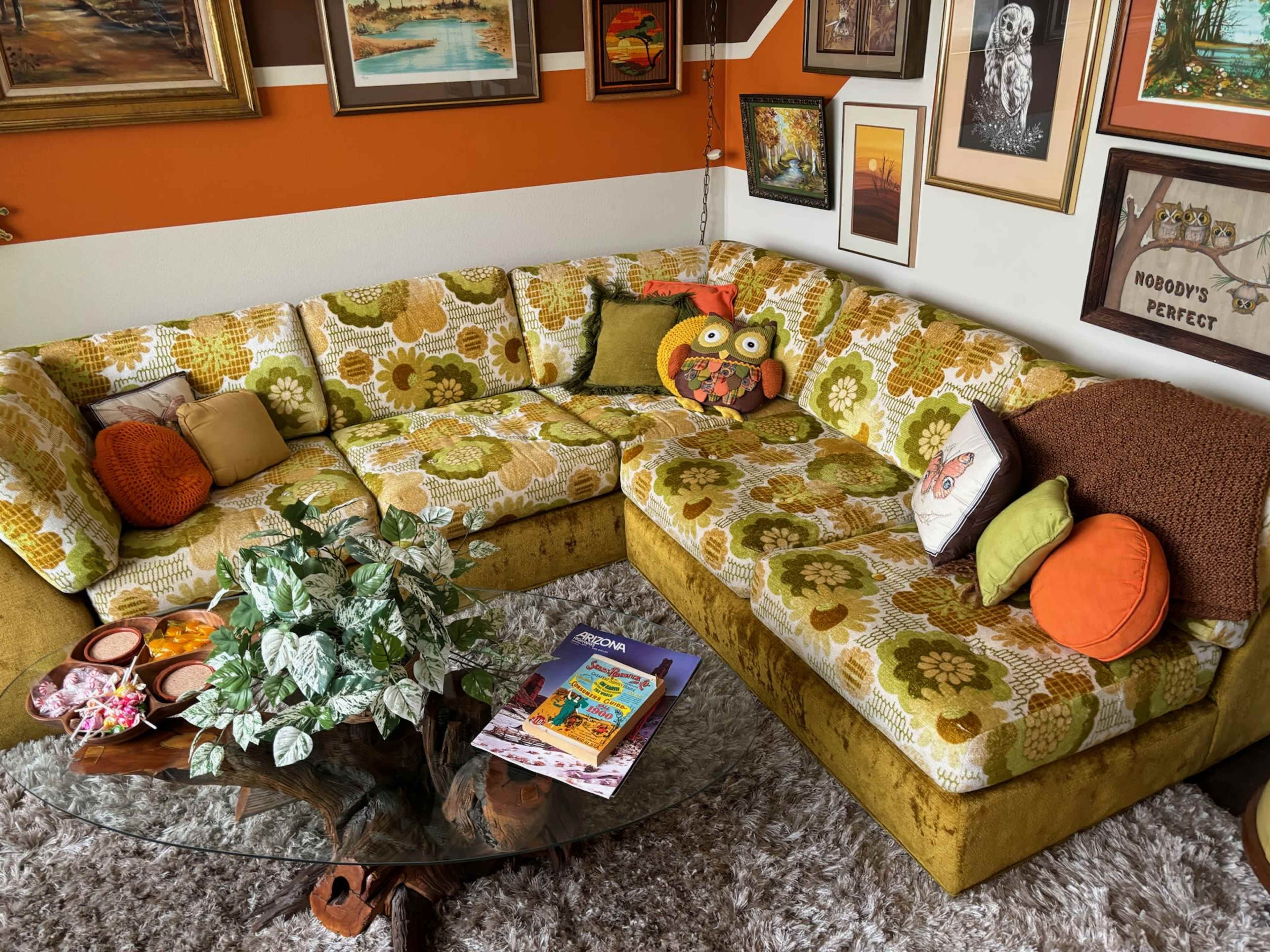 A vintage-style living room with a large, patterned sectional sofa, a glass coffee table, and various framed artworks on the walls.