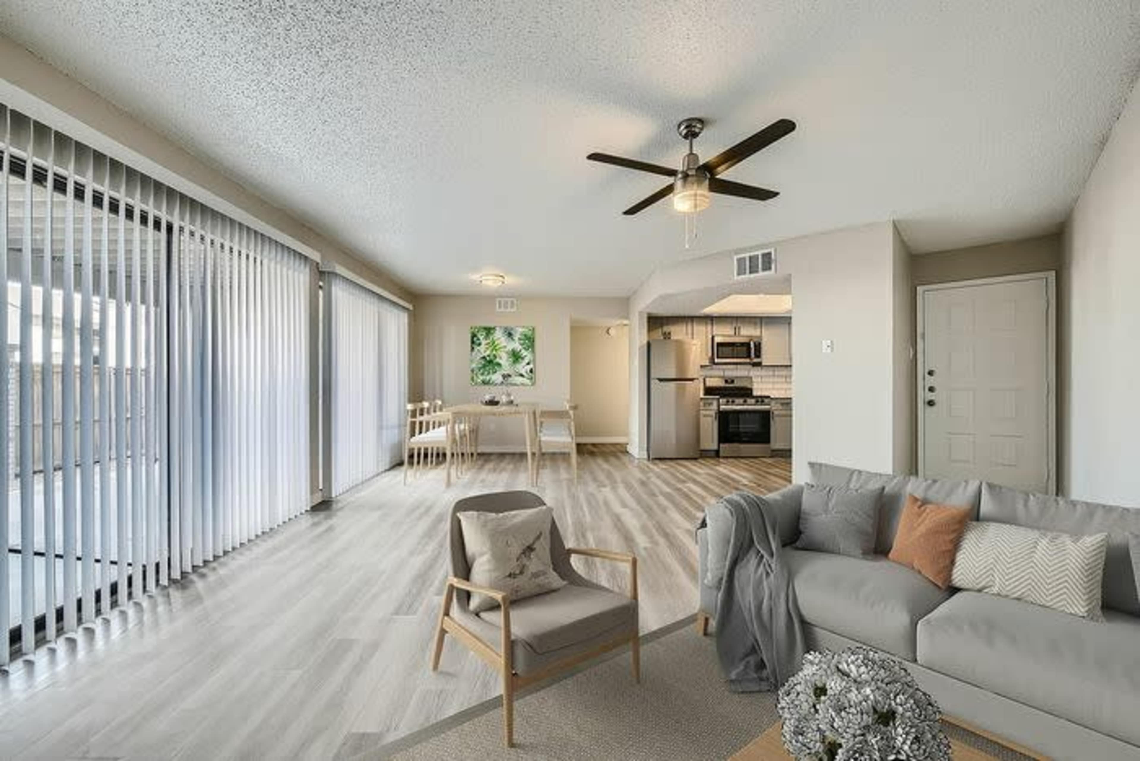 The image shows a modern living area with an open layout featuring a sofa, a chair, and a dining area visible in the background, along with sliding glass doors leading to a balcony.