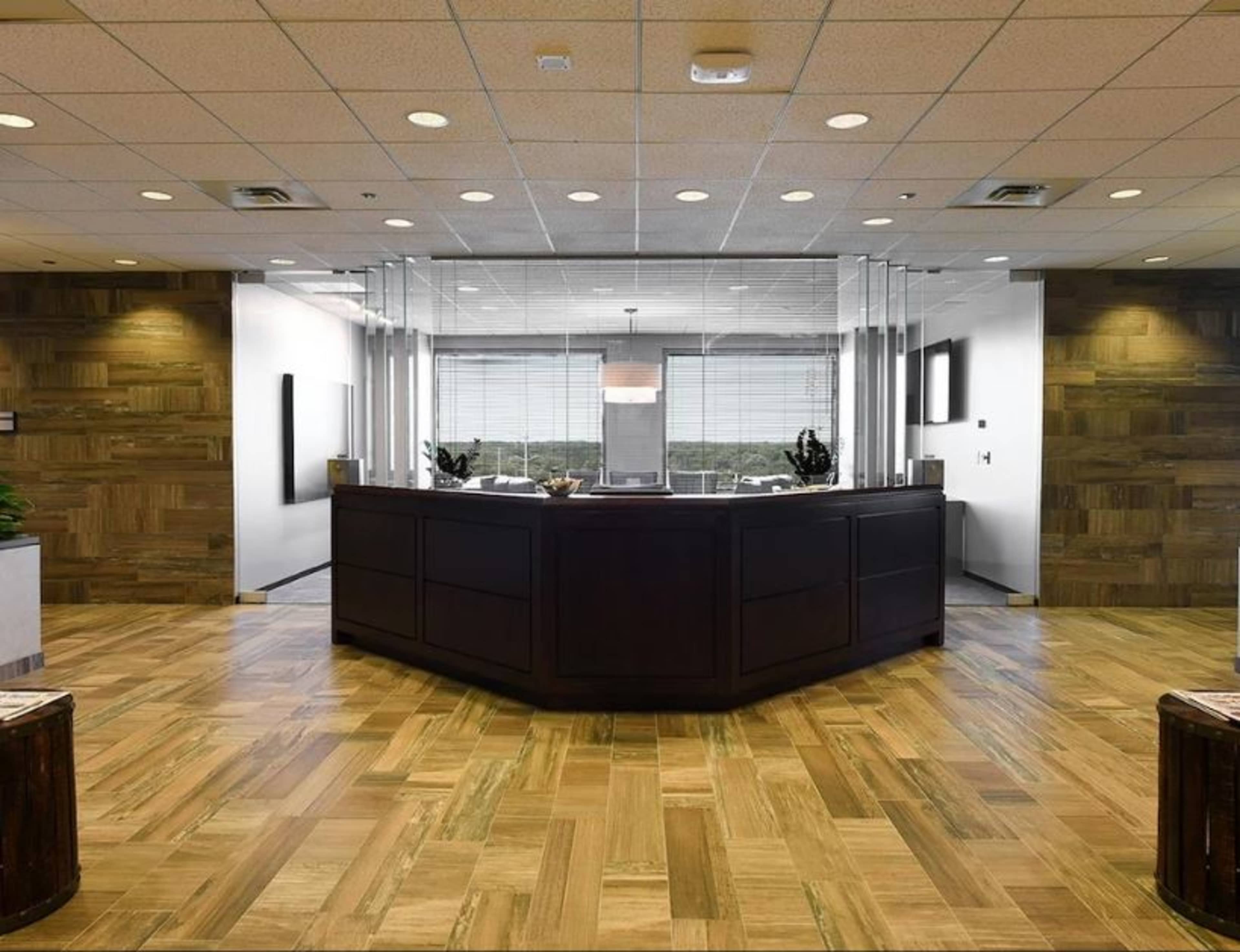 A modern office reception area with a dark wooden front desk, large windows, and a wooden floor.