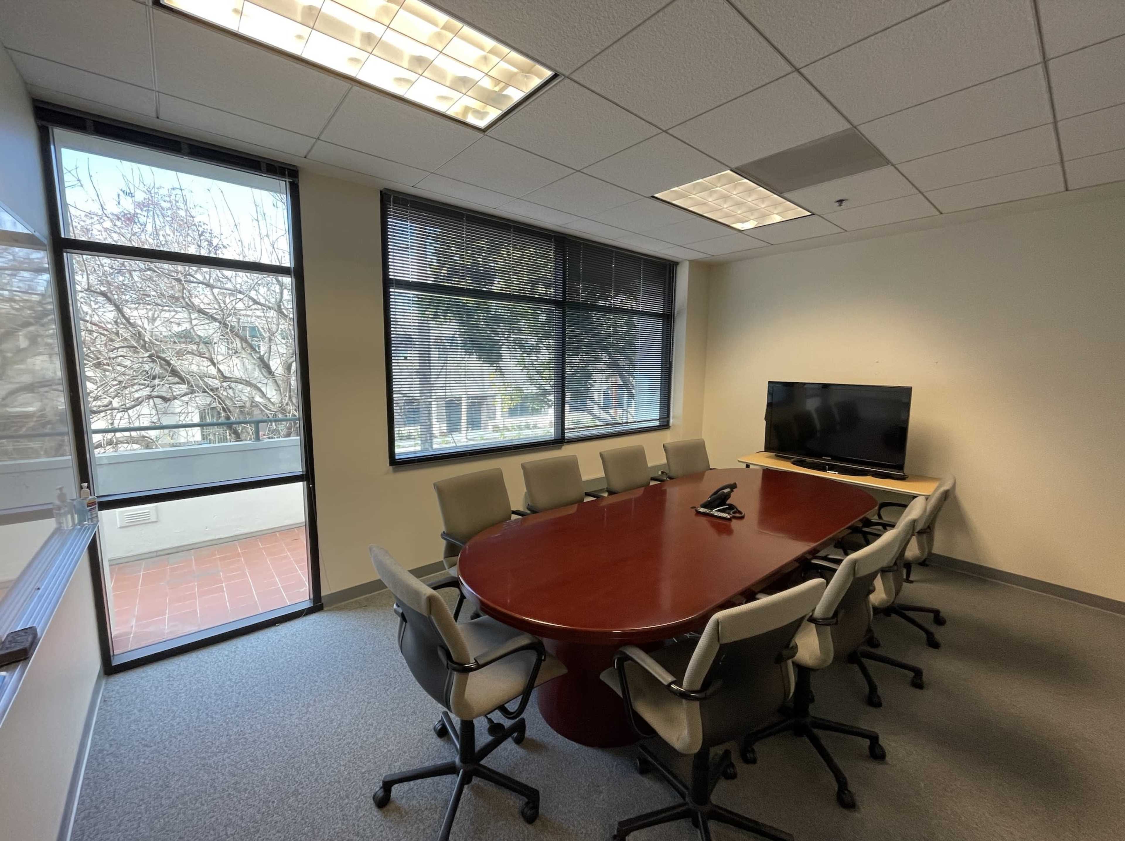 A conference room features a large wooden oval table surrounded by eight chairs, with a wall-mounted television and large windows.