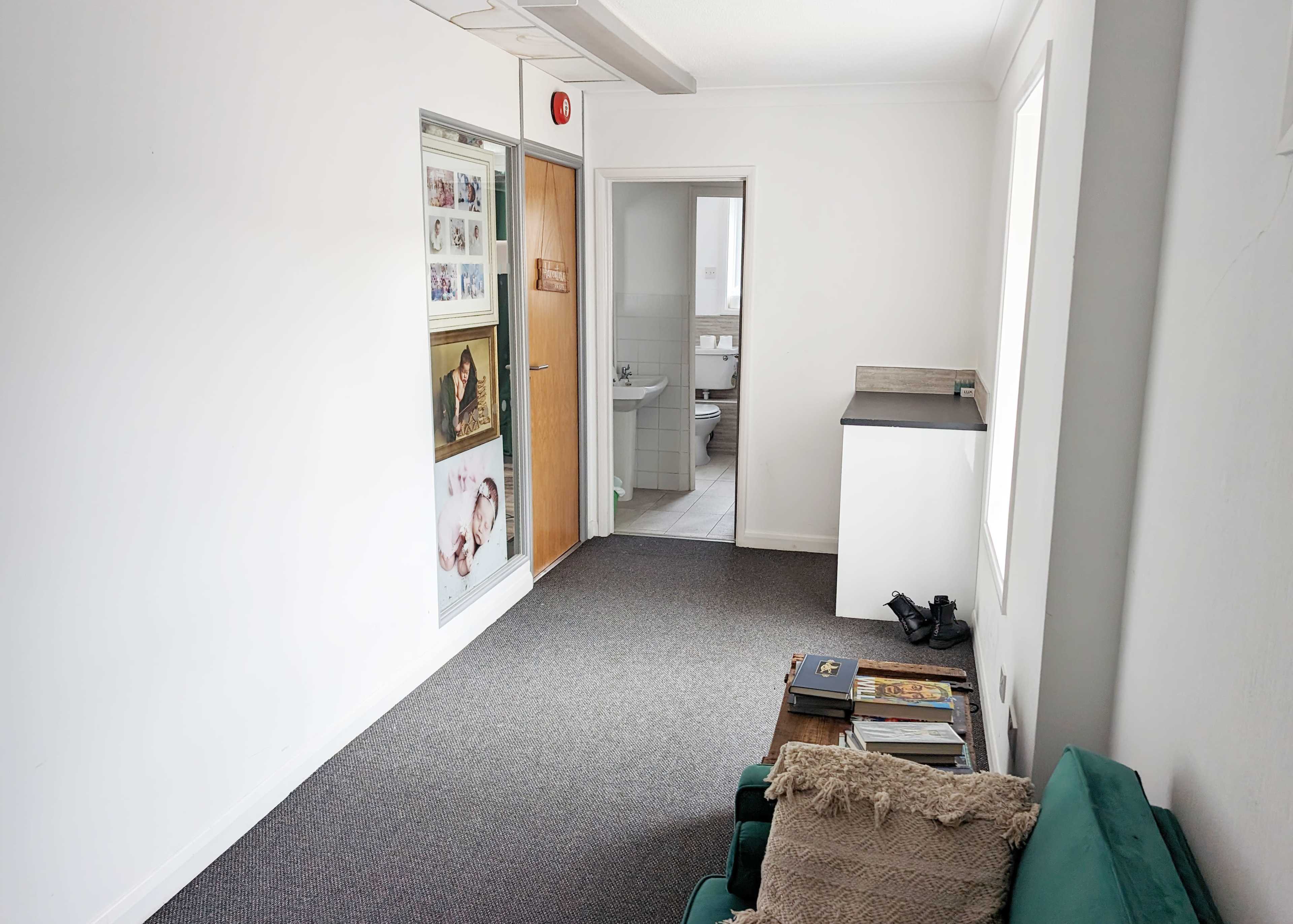 A small, open room with white walls, a carpeted floor, and a green couch, featuring a table with magazines and a door leading to a bathroom in the background.