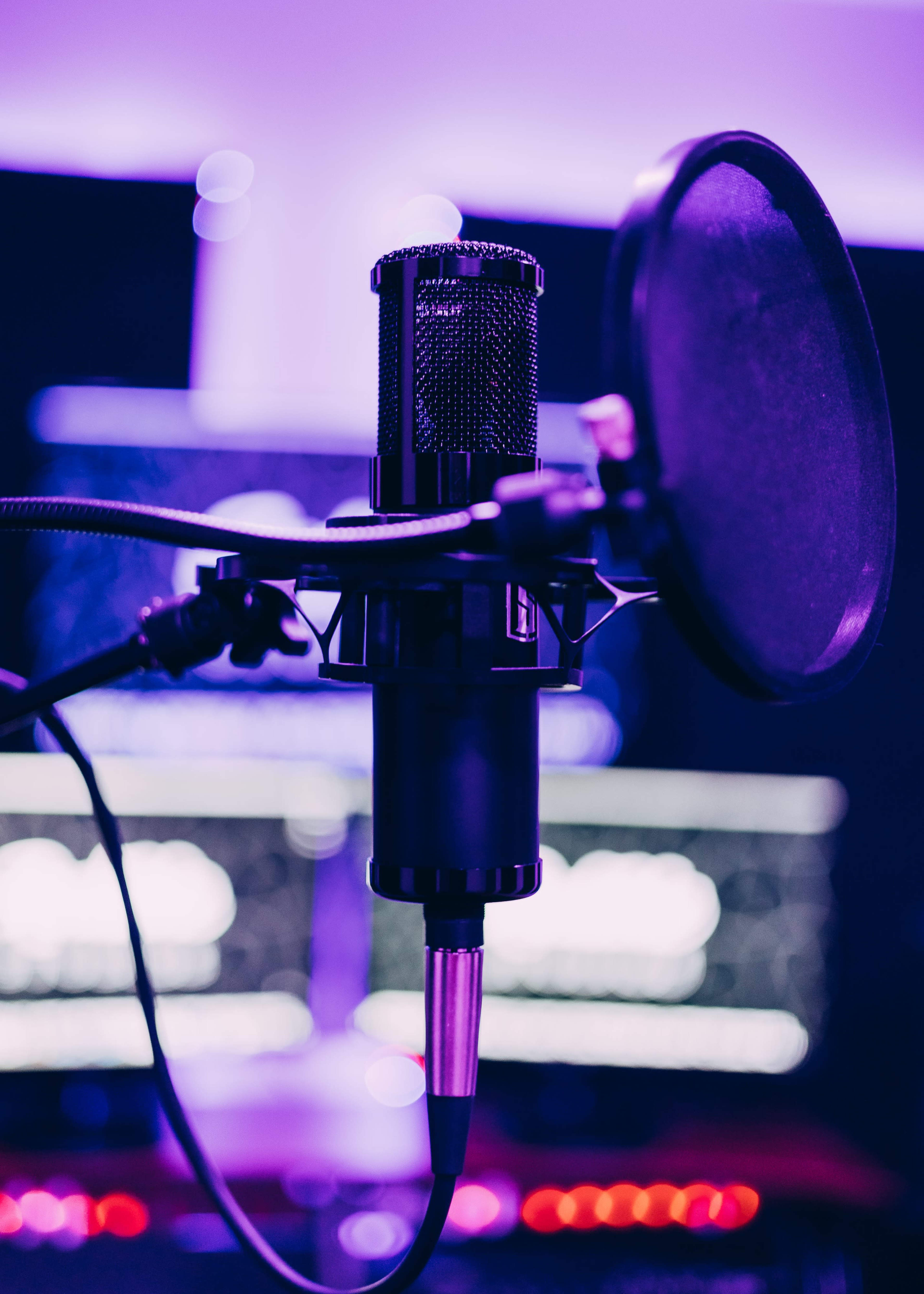 A microphone with a pop filter is positioned in front of computer screens illuminated by purple lighting.