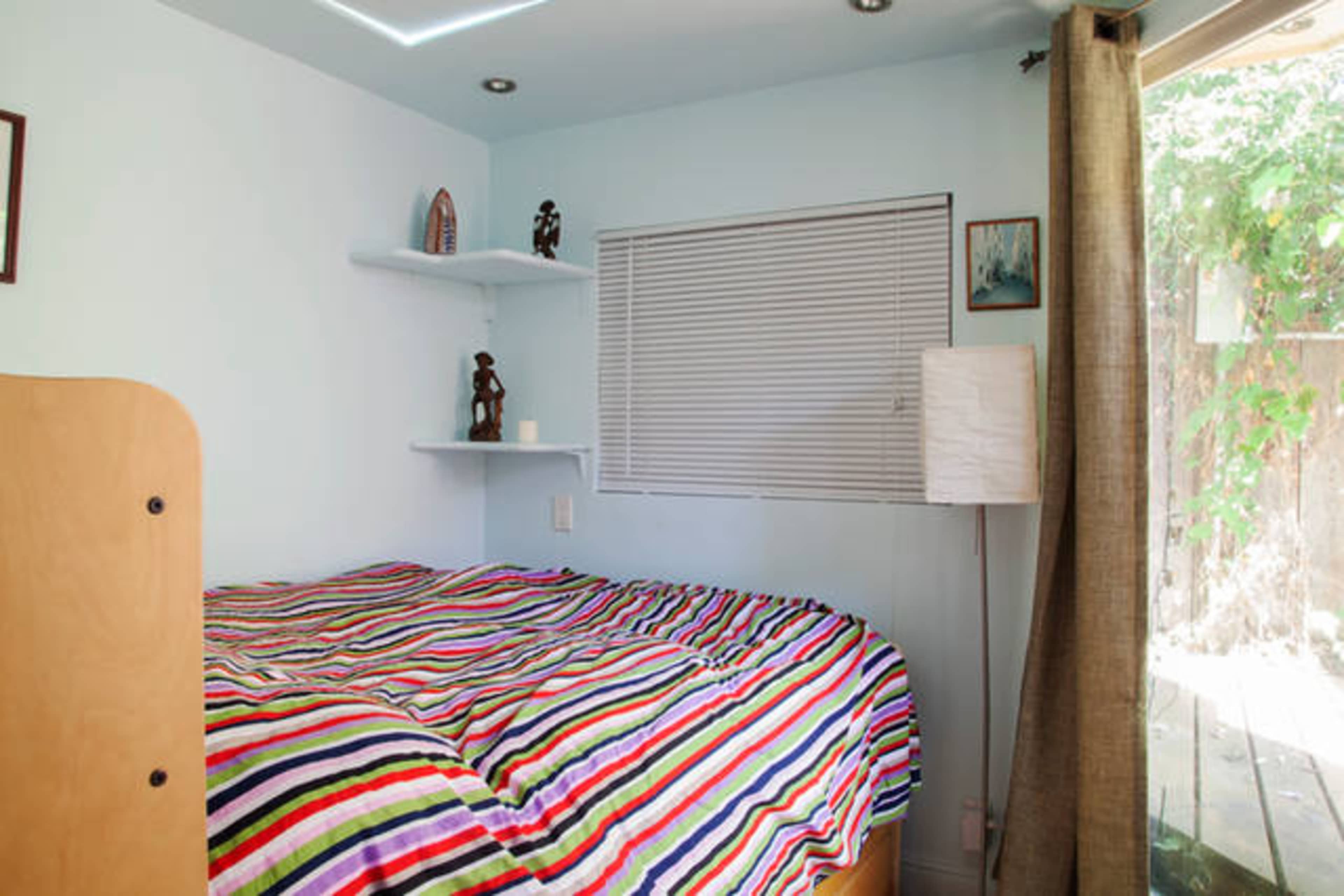 A bedroom with a striped comforter on a bed, shelves on the wall, a floor lamp, and a window with curtains.