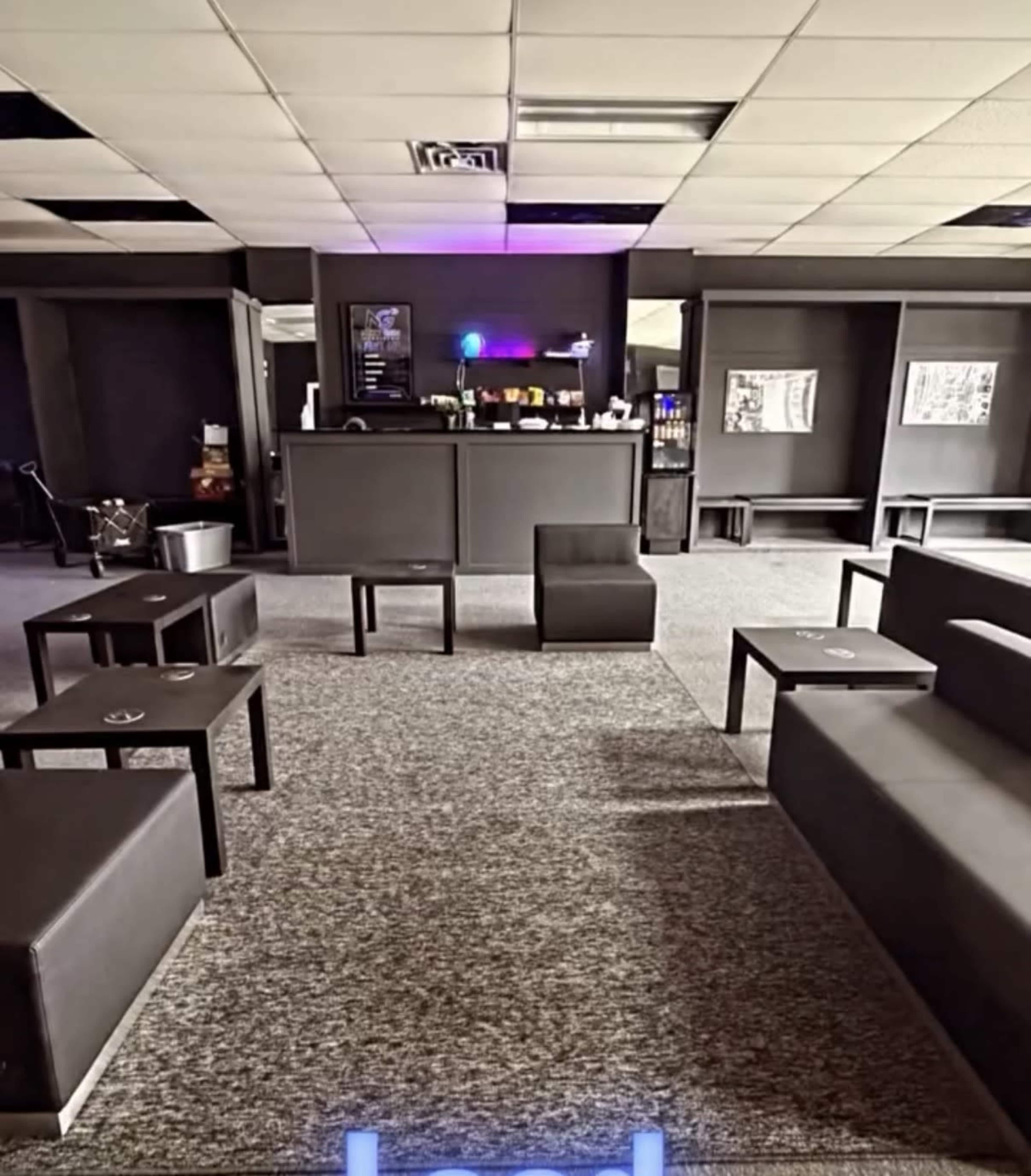 The image shows a modern lounge area with dark furniture, a bar counter, and gray carpeting.