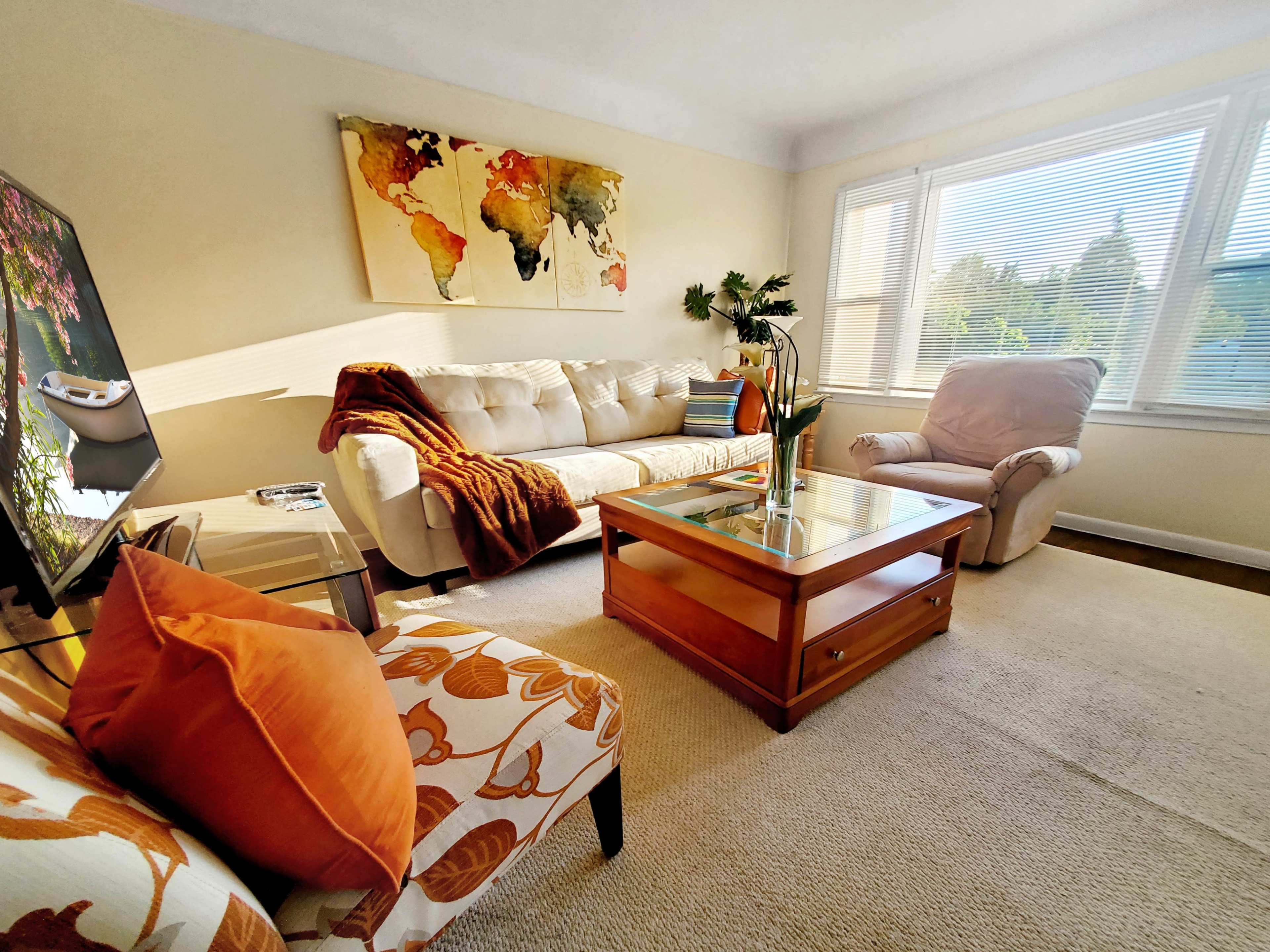 A cozy living room features a beige sofa, a glass-topped coffee table, and an armchair, with a world map artwork on the wall and sunlight streaming through the windows.