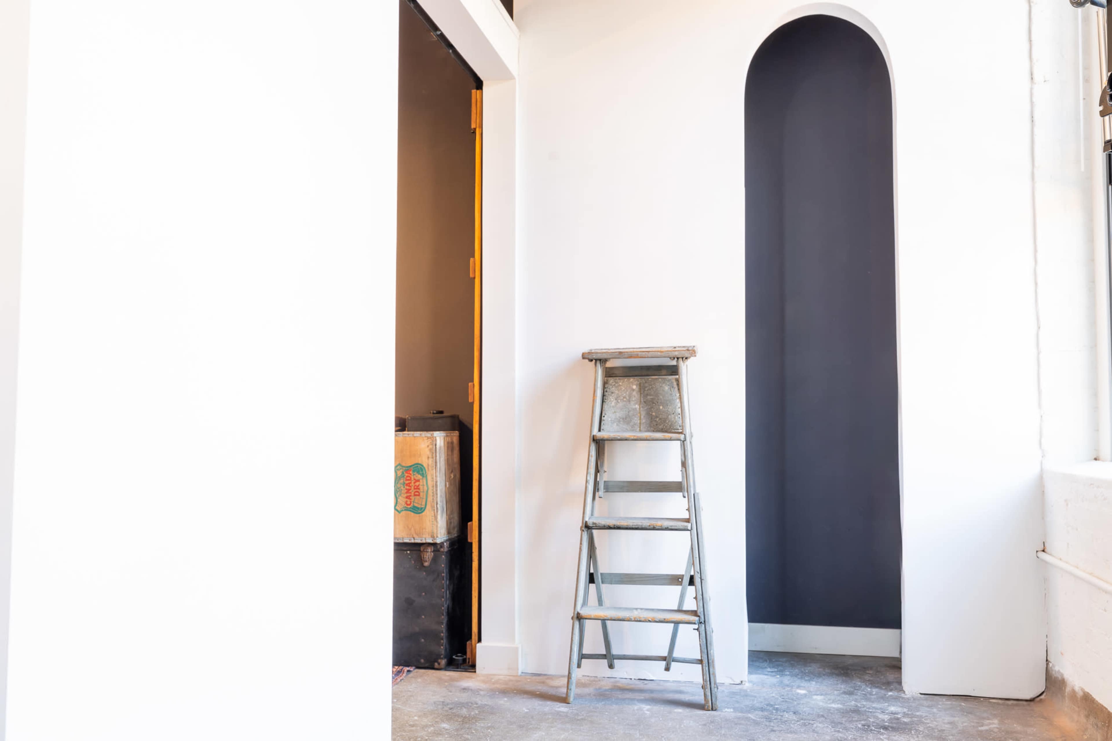 A metal step ladder stands next to a wall featuring an arched recess, with a dark-colored section contrasting a white wall.
