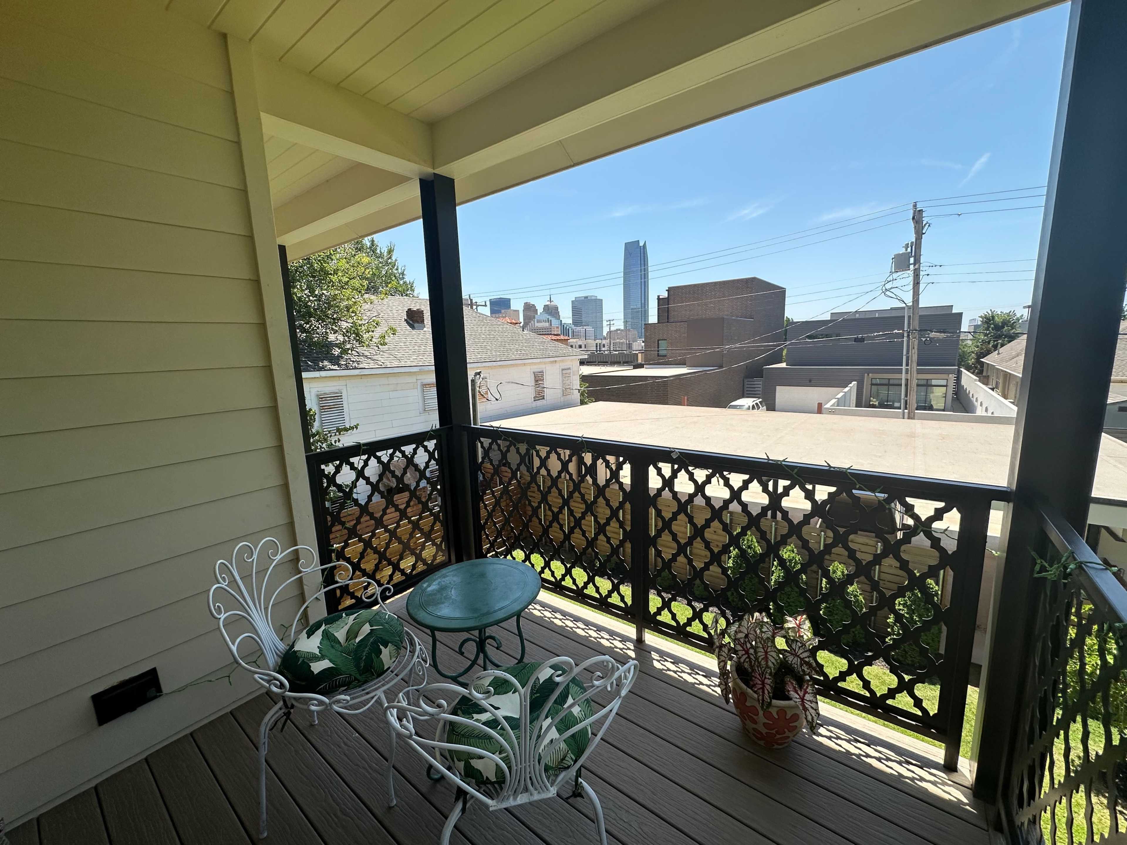 A small balcony with two white metal chairs and a round green table, overlooking a city skyline in the distance.