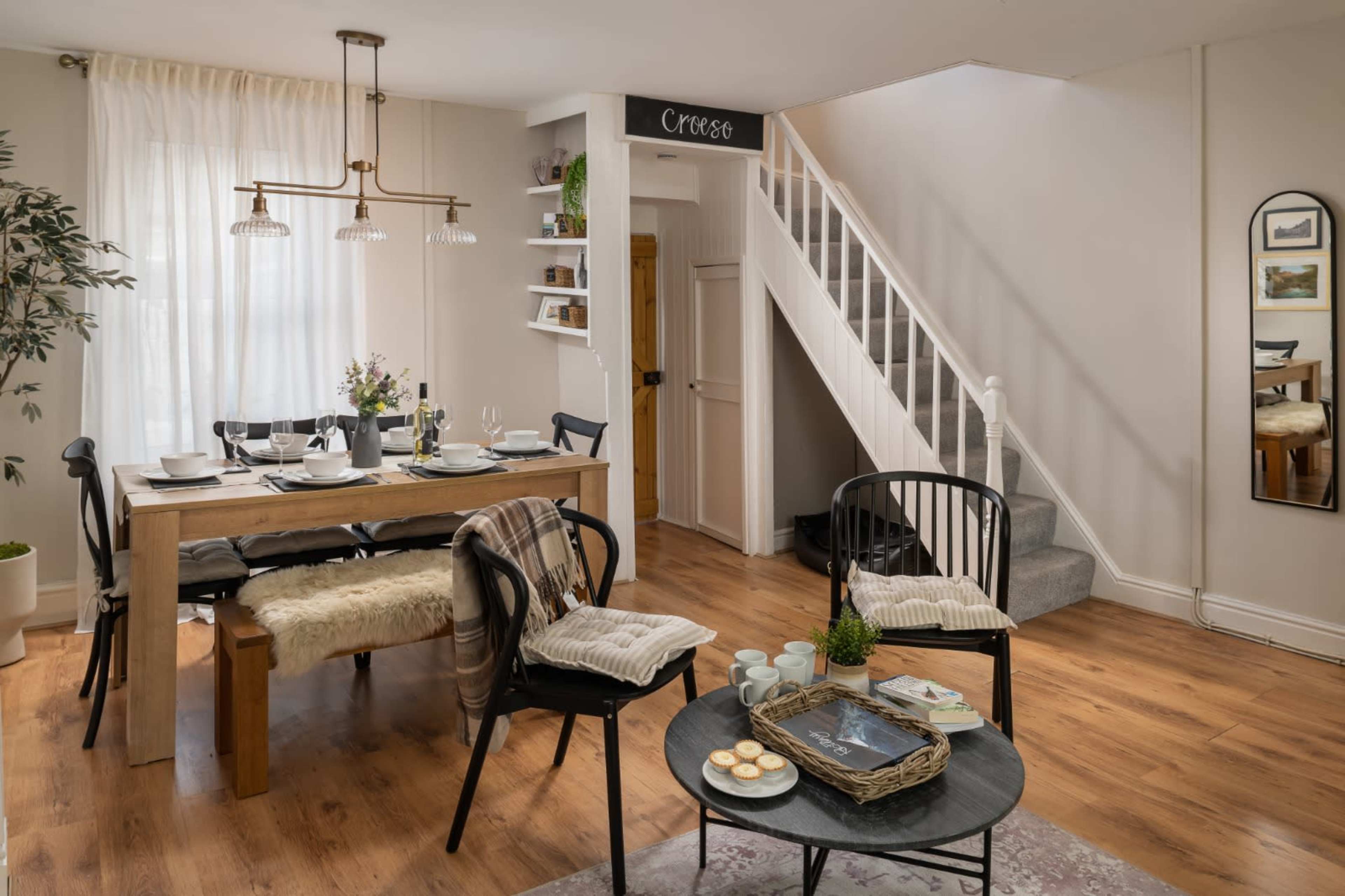 The image shows a dining area with a wooden table set for a meal, a staircase in the background, and a cozy seating arrangement nearby.