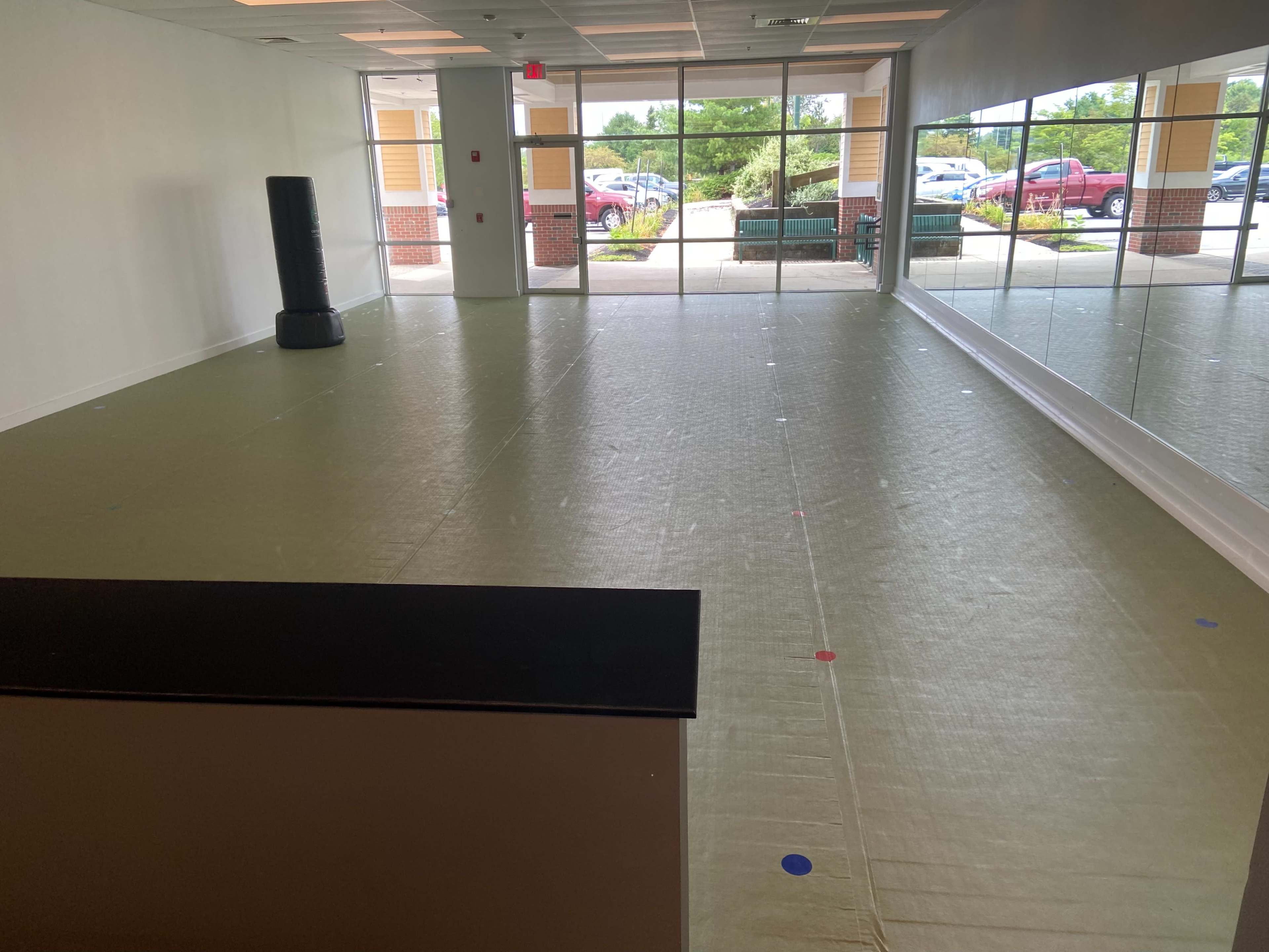An empty room with a smooth, light-colored floor, a mirrored wall on one side, and large windows letting in natural light.