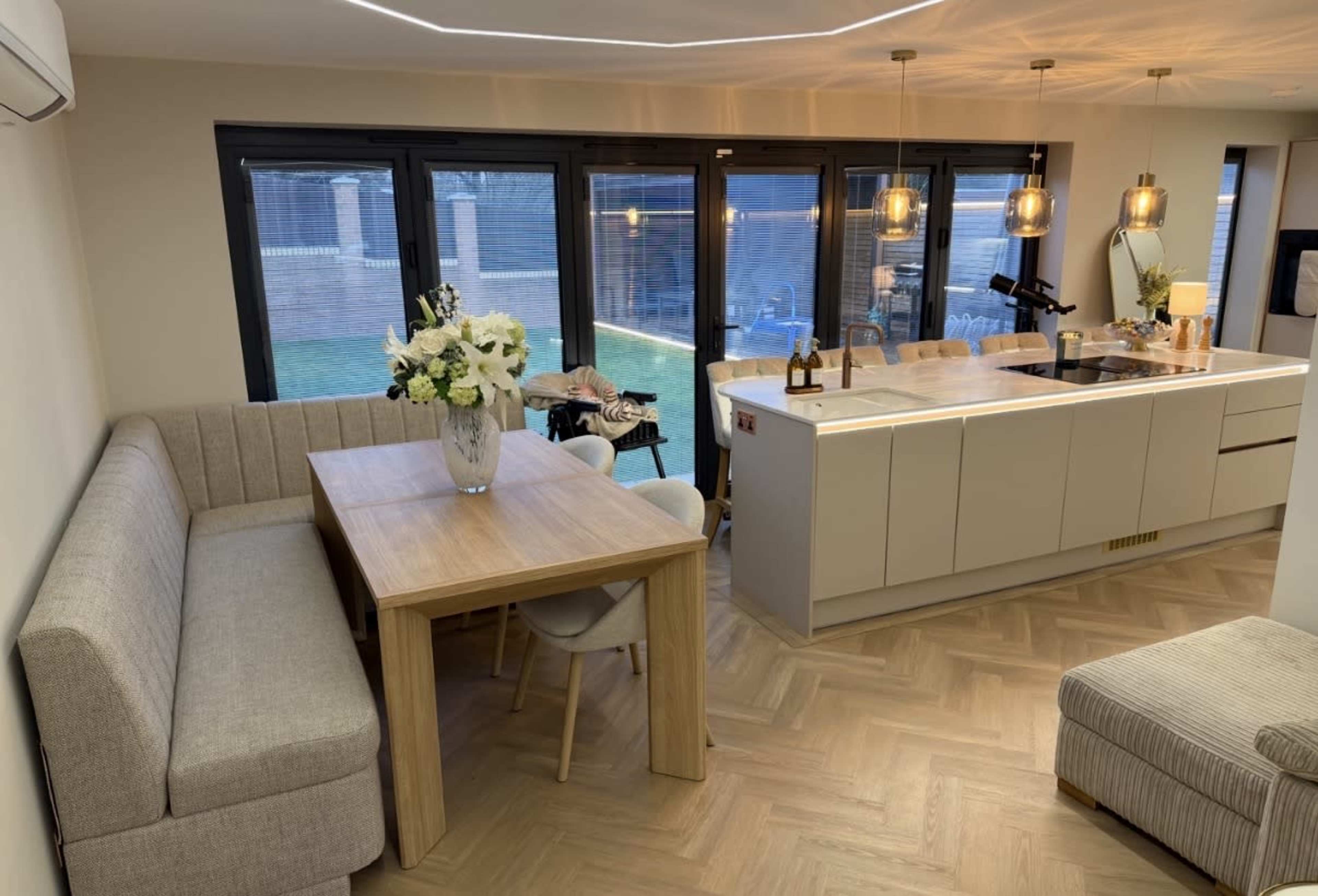 The image shows a modern open-concept kitchen and dining area featuring a light wood dining table, a gray upholstered bench, and large windows overlooking a backyard.