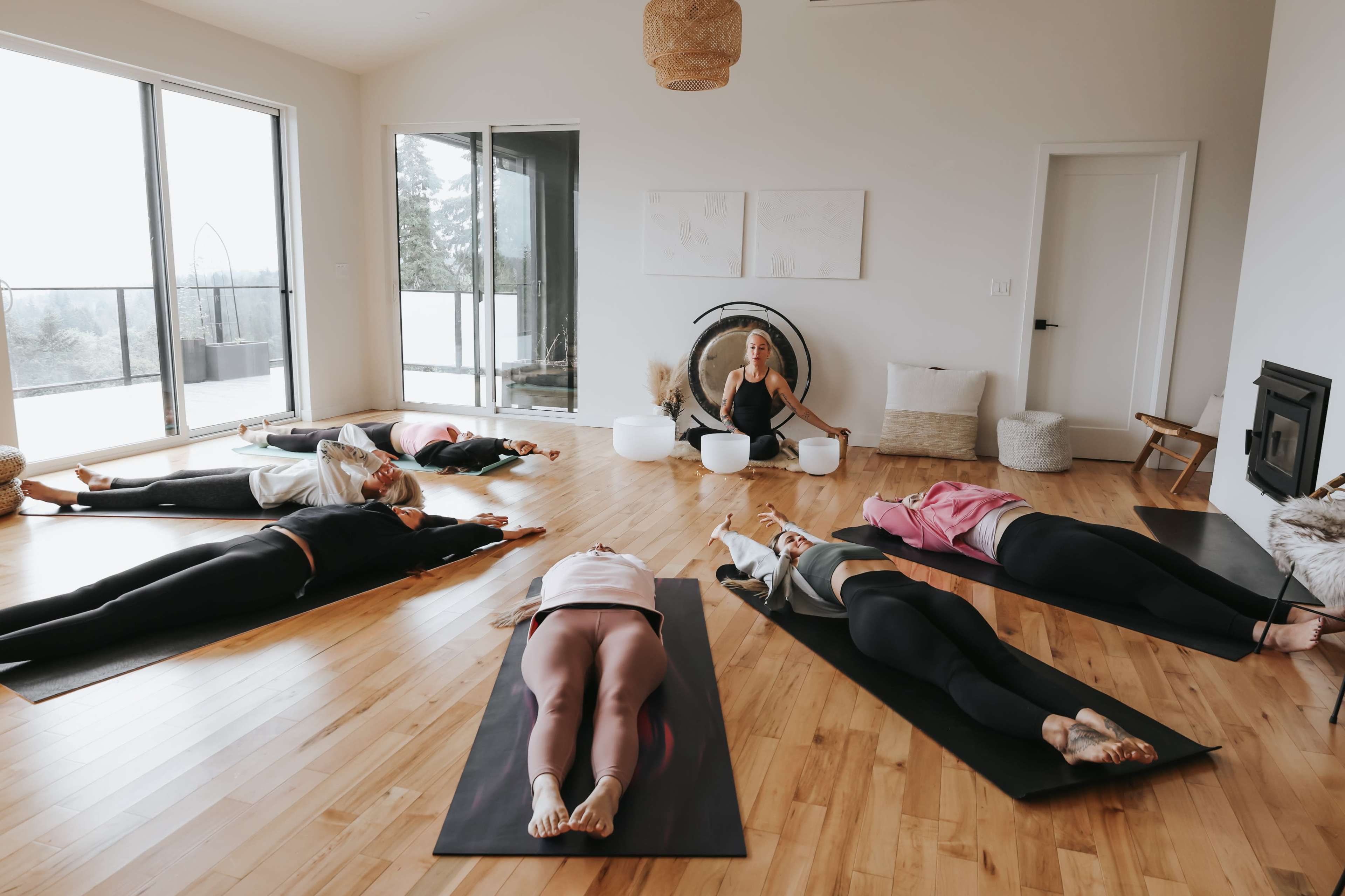 A group of individuals lies on yoga mats in a spacious, bright room while a person at the front leads a session.
