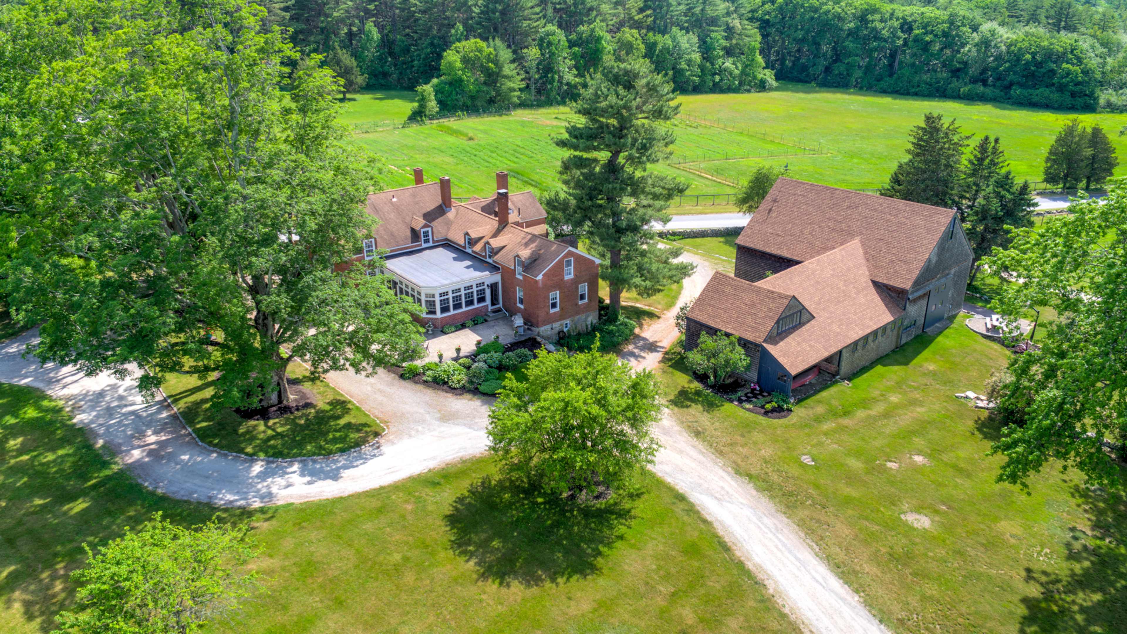 The aerial view shows a large property featuring a red-brick house with a sunroom, a barn, and lush green fields surrounding them.