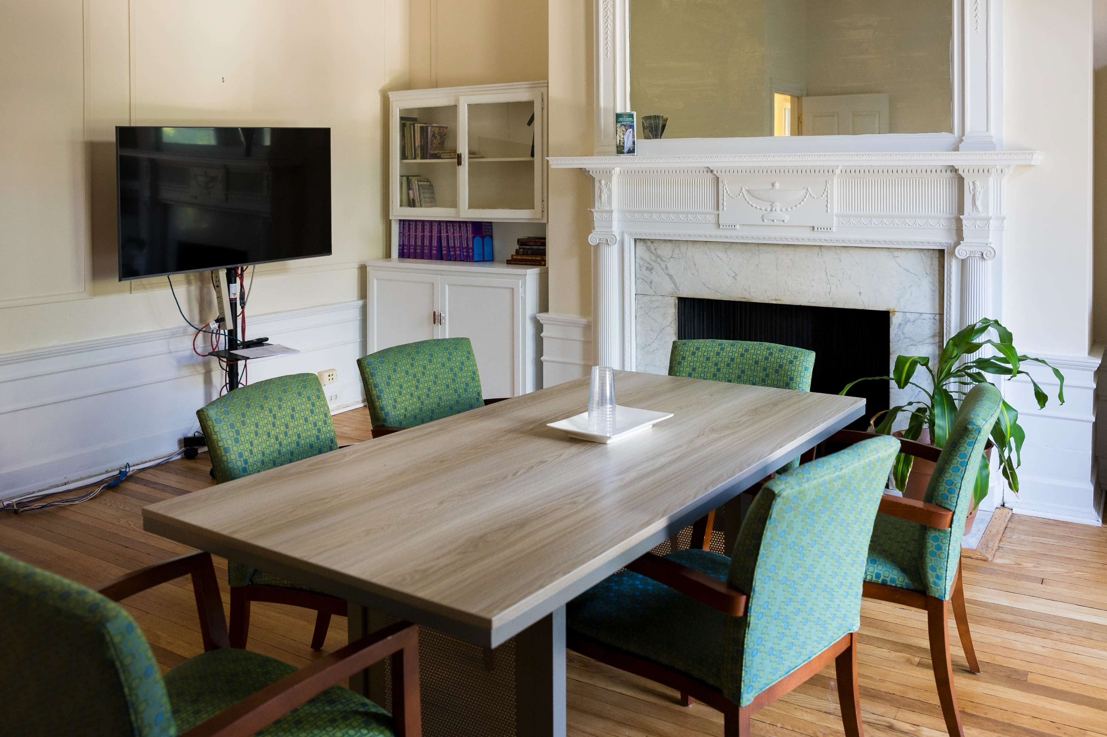 A spacious room features a wooden table surrounded by green upholstered chairs, a television mounted on the wall, and a decorative fireplace.