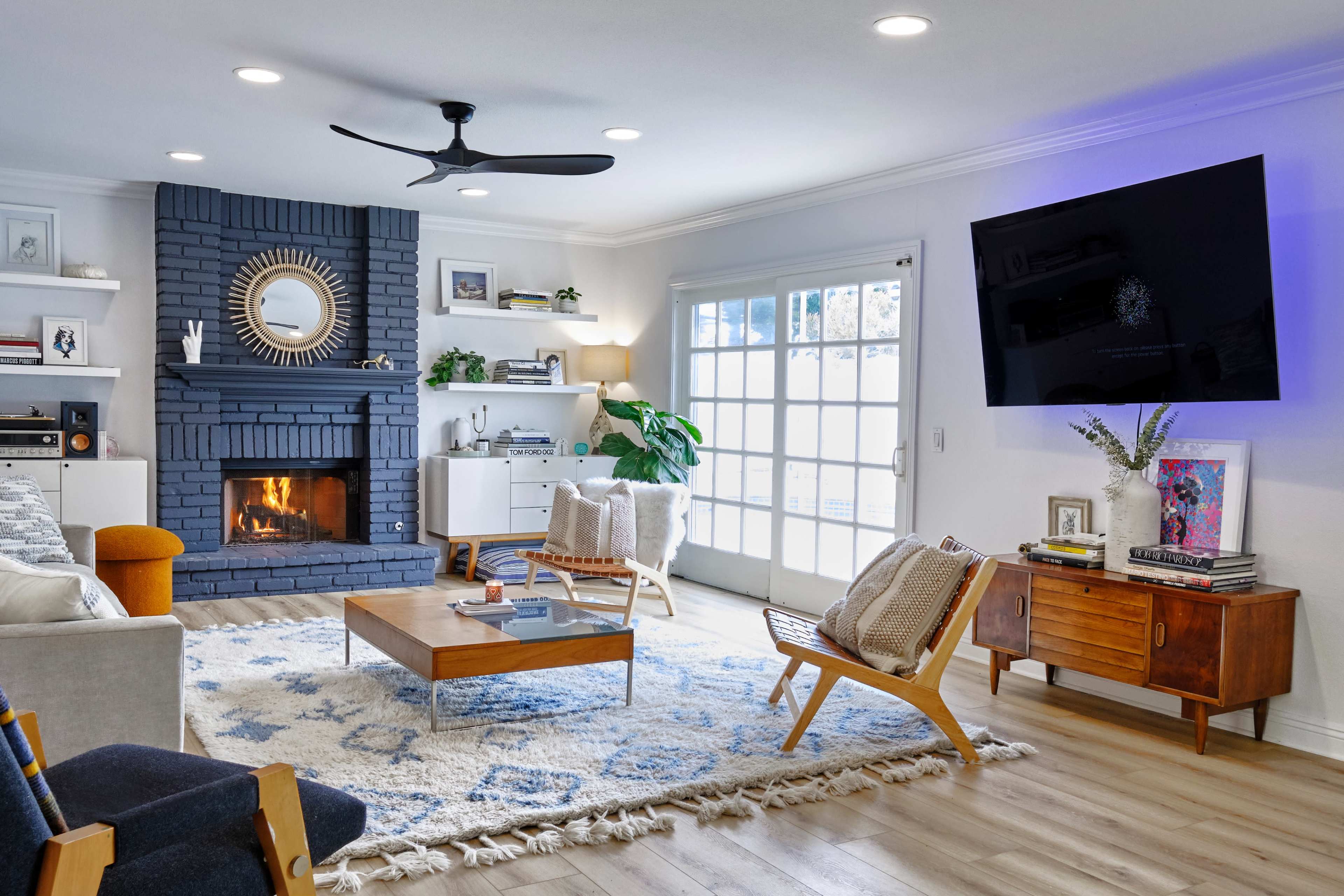 A modern living room features a brick fireplace, a large wall-mounted TV, comfortable seating, and a wooden coffee table on a patterned rug.
