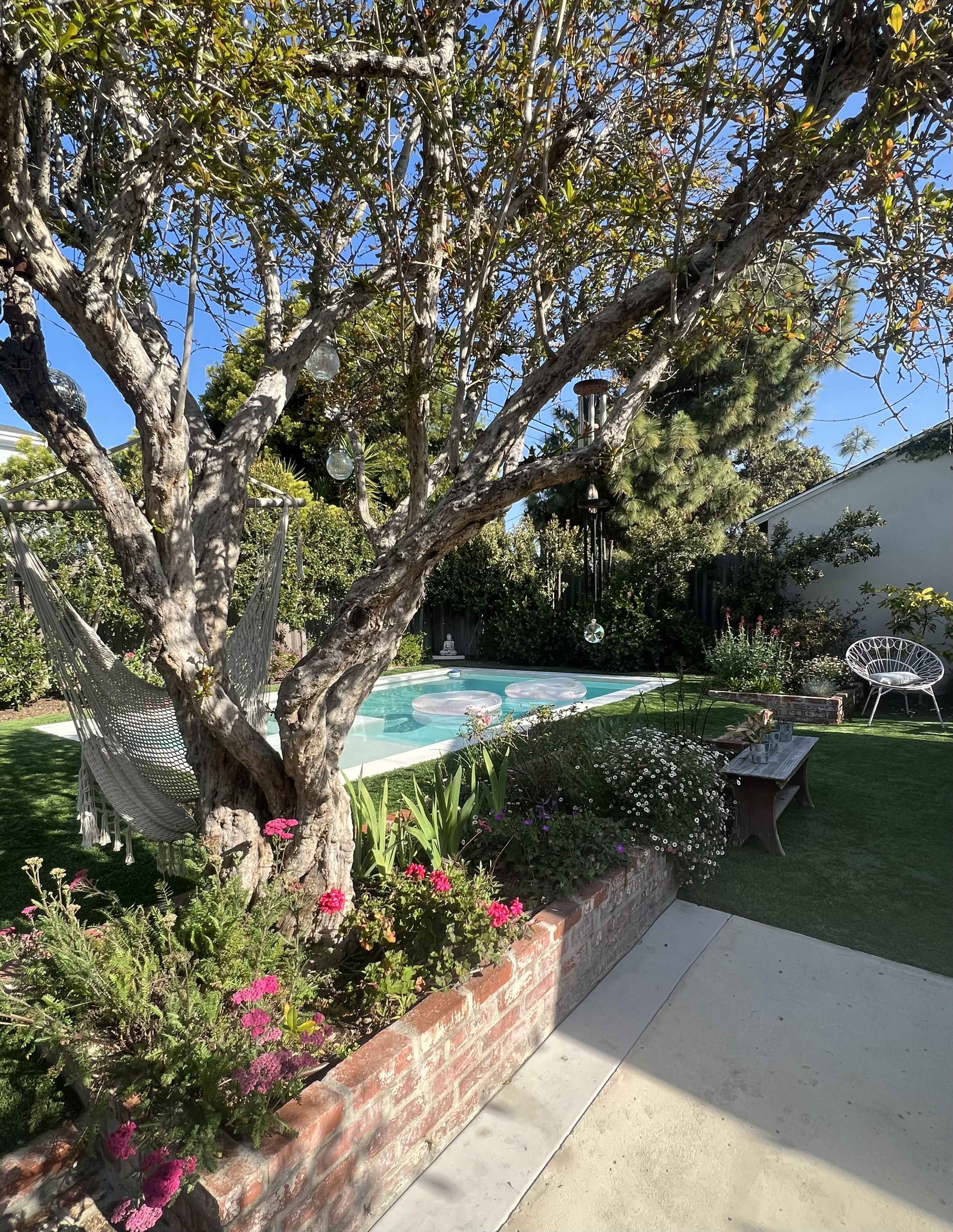 A backyard with a swimming pool surrounded by gardens, a hammock hanging from a tree, and a brick planter filled with flowers.